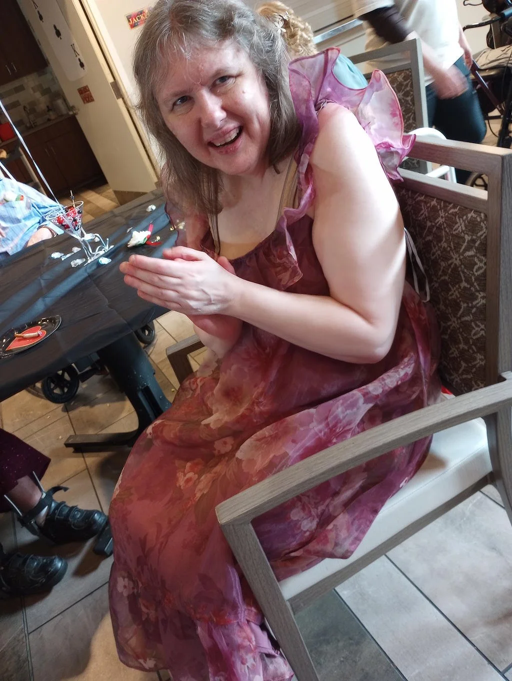 A woman with gray hair and fair skin, wearing a pink and floral dress with sheer ruffled sleeves, is sitting on a chair at a table. She is smiling with her hands pressed together in a prayer position. The setting appears to be a dining area with othe