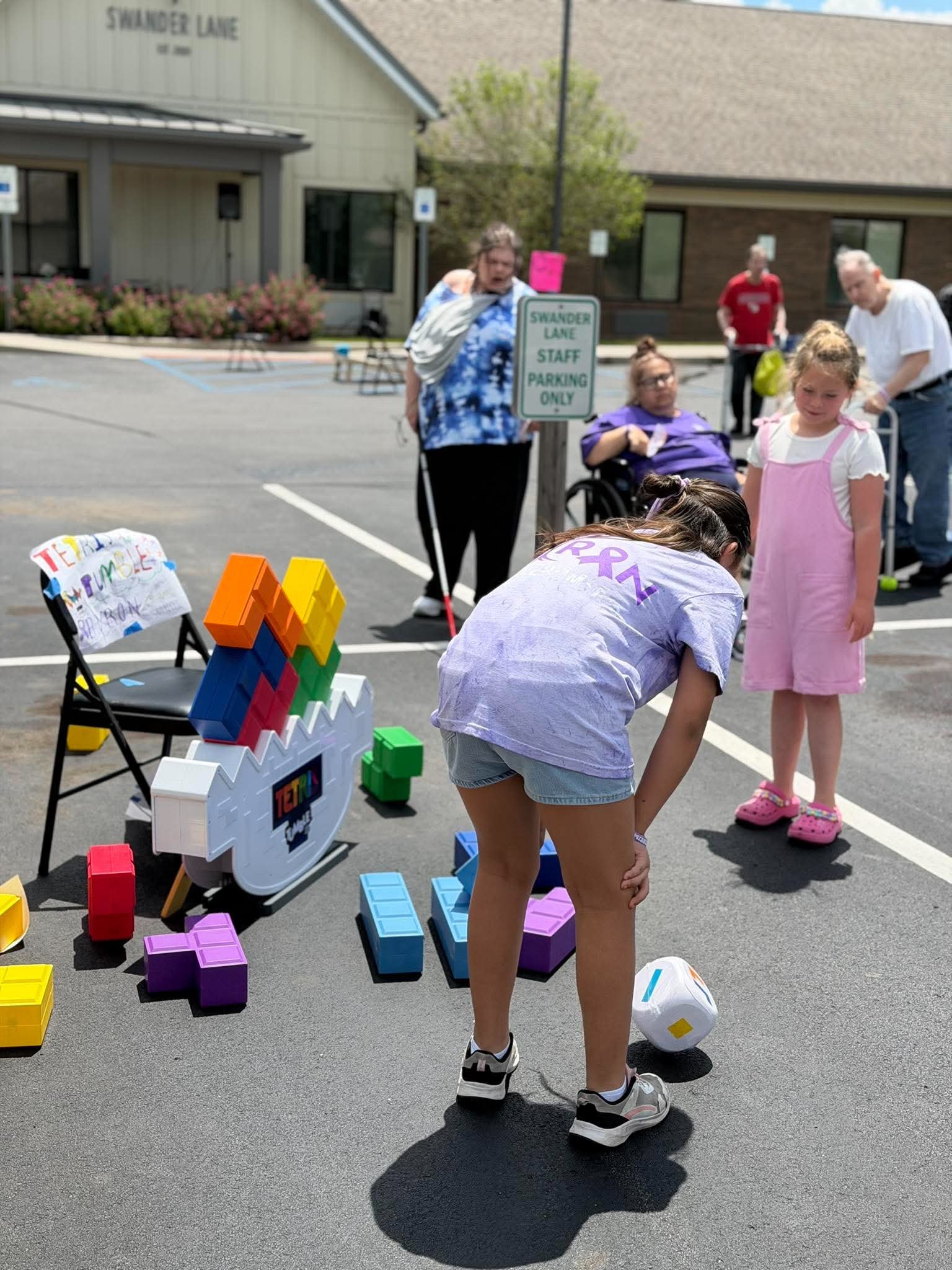 Children playing with colorful building blocks on a parking lot, with adults supervising, near a building with a sign that reads "Shander Lane Staff Parking Only."