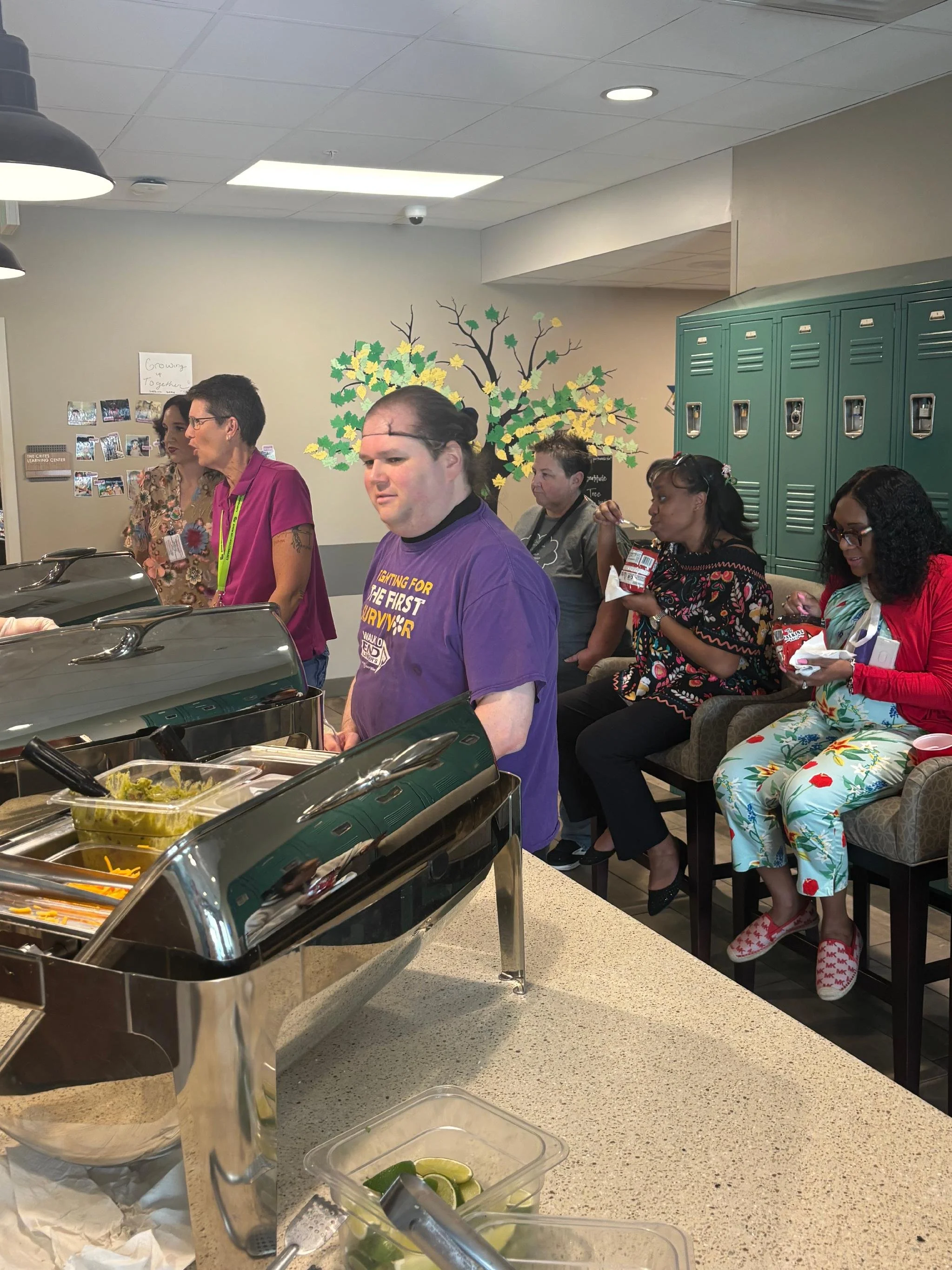 People seated in a waiting area near a buffet, some eating snacks, with lockers and a wall mural of a tree in the background.