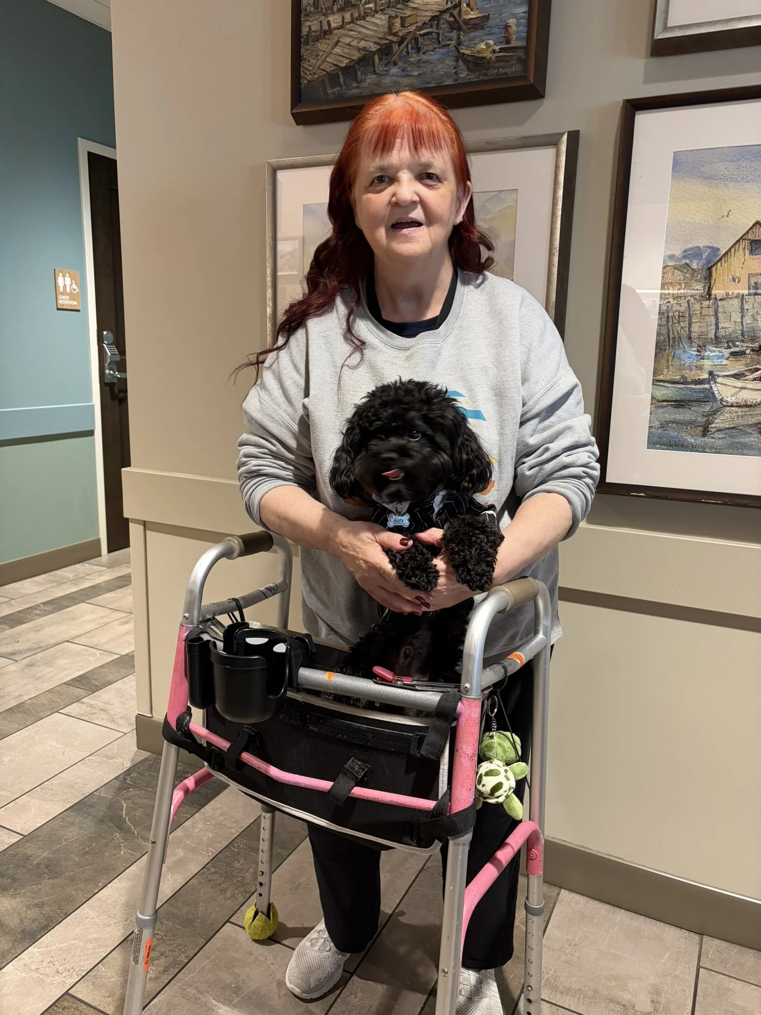 A woman with red hair smiling and holding a small black puppy sitting in a walker in an indoor setting with framed artwork on the wall behind her.