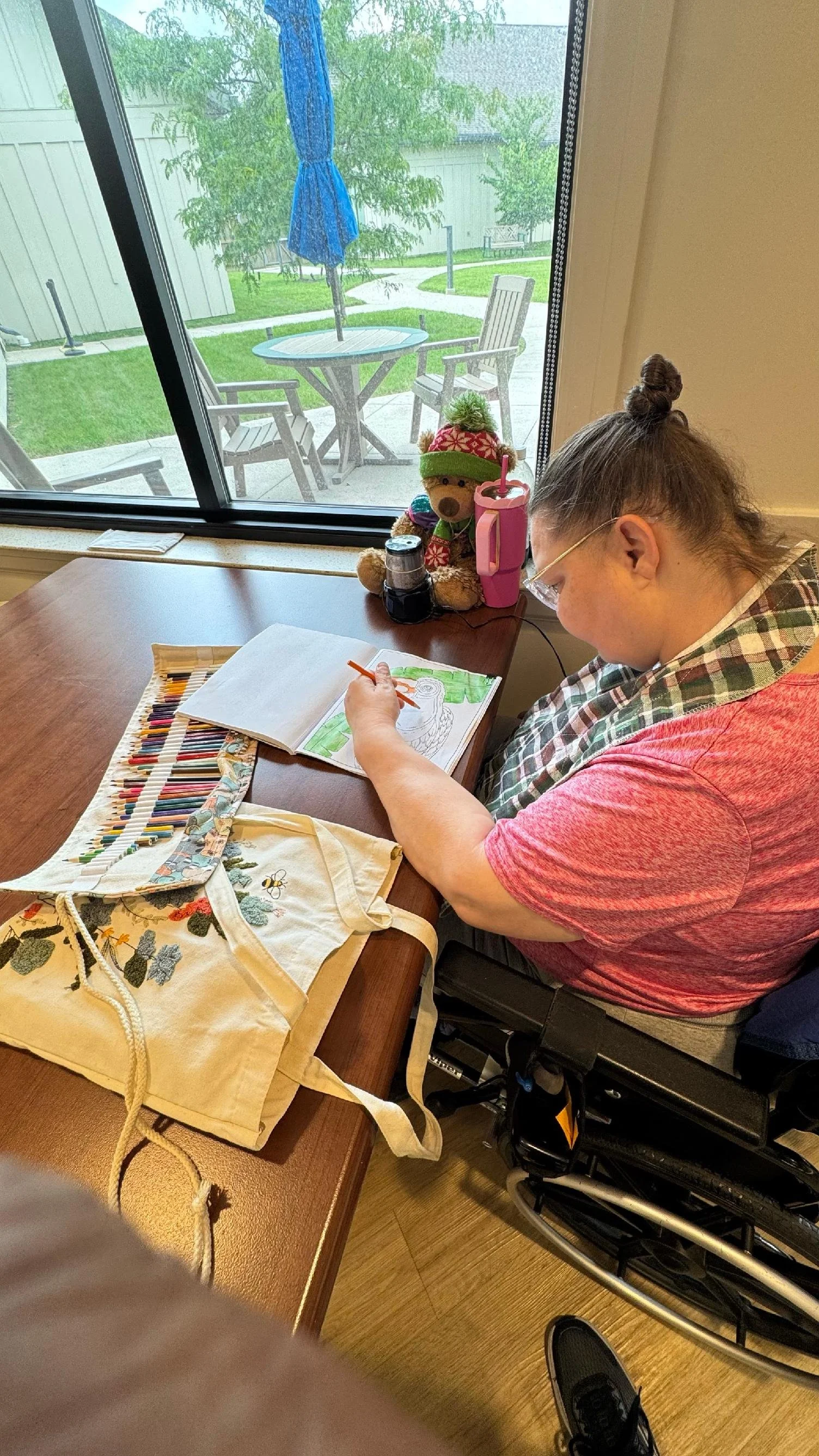 A woman in a wheelchair coloring in a book at a wooden table near a window, with a tote bag, teddy bear, and art supplies on the table. Outside, there is a patio with chairs, a table with an umbrella, and a lawn with trees.