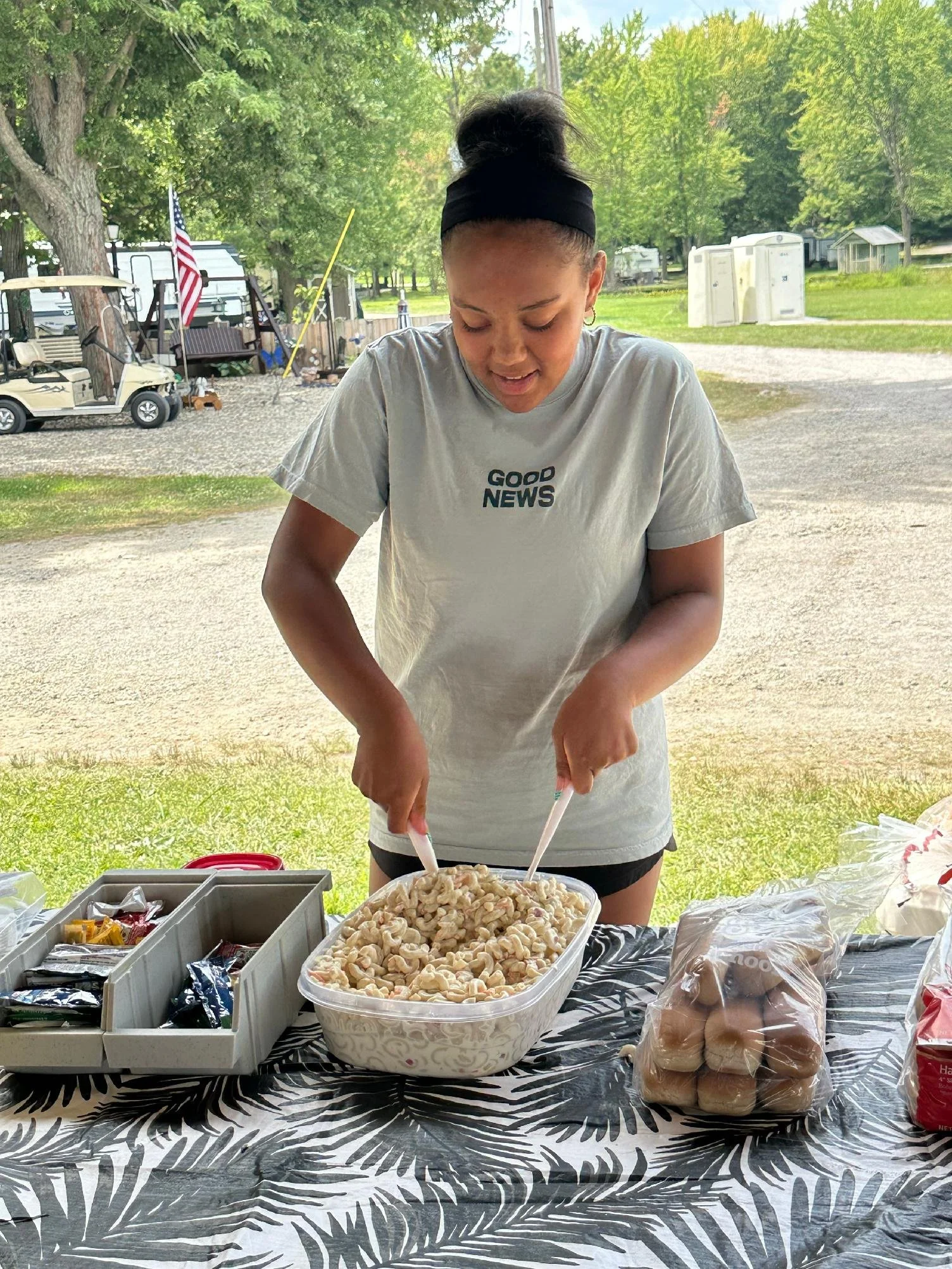 A woman wearing a gray t-shirt with the words 'Good News' on it, standing outdoors at a table, shucking corn into a large bowl. The table has a black and white patterned cloth and various food items. Behind her, there are green trees, a lawn, a grave
