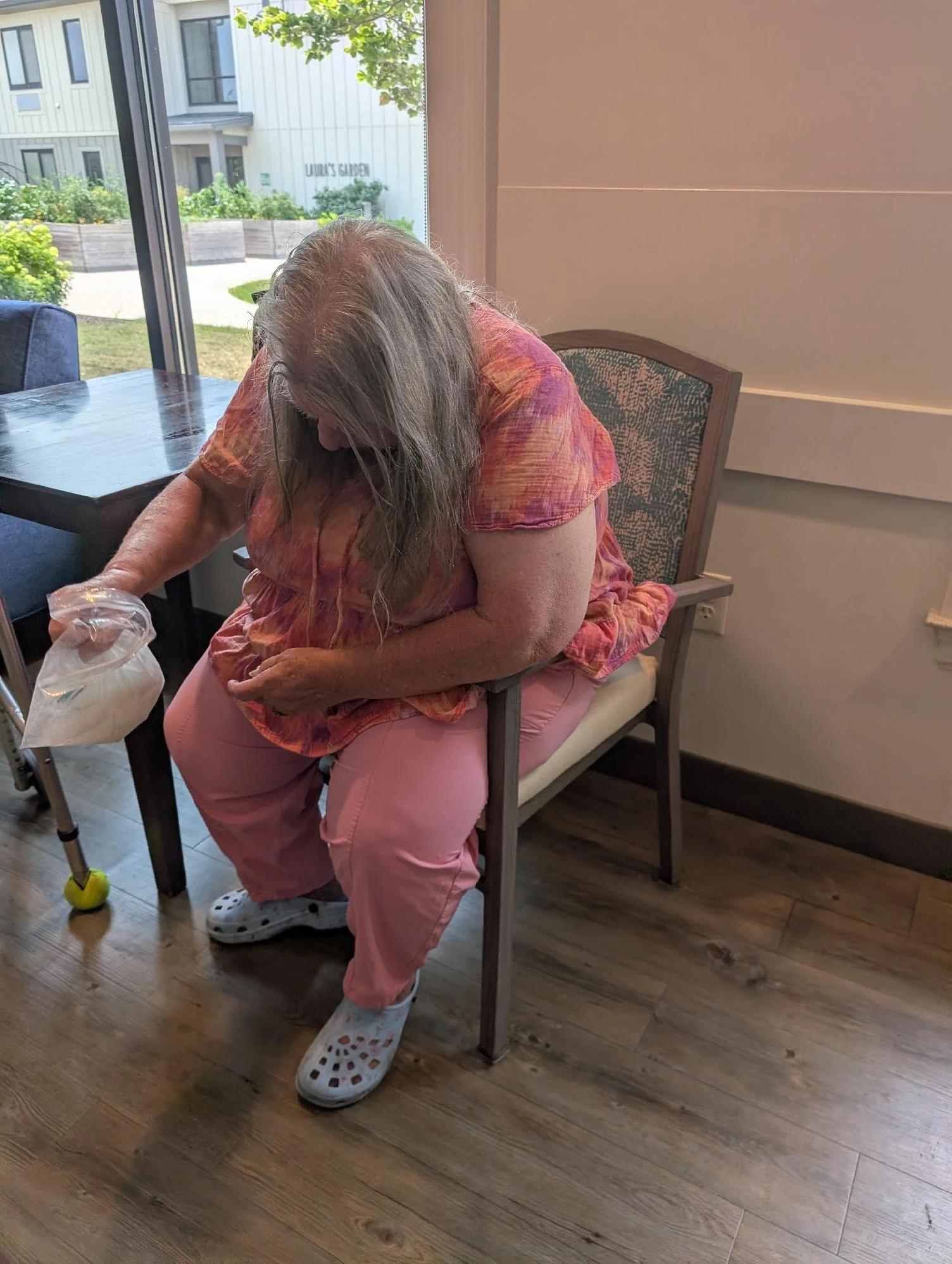 An elderly woman with long gray hair sitting on a chair at a table inside a cafe or restaurant, leaning forward and looking down at a plastic bag in her hand, wearing a colorful shirt, pink pants, and white Crocs with holes. A window behind her shows