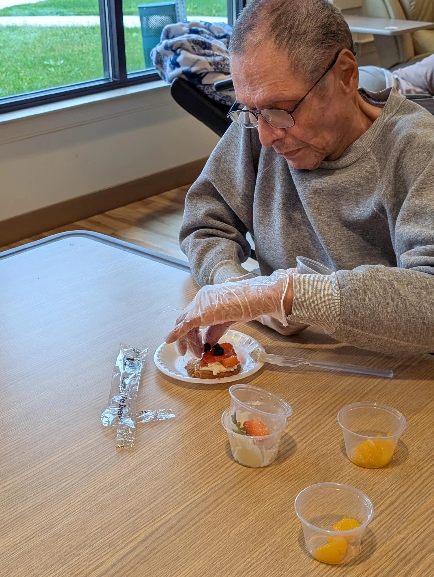 An elderly man wearing glasses and a gray sweatshirt is sitting at a wooden table with a dessert on a paper plate. The dessert appears to be a pastry with fruit on top. There are small cups with fruit toppings or garnishes on the table. The man is we
