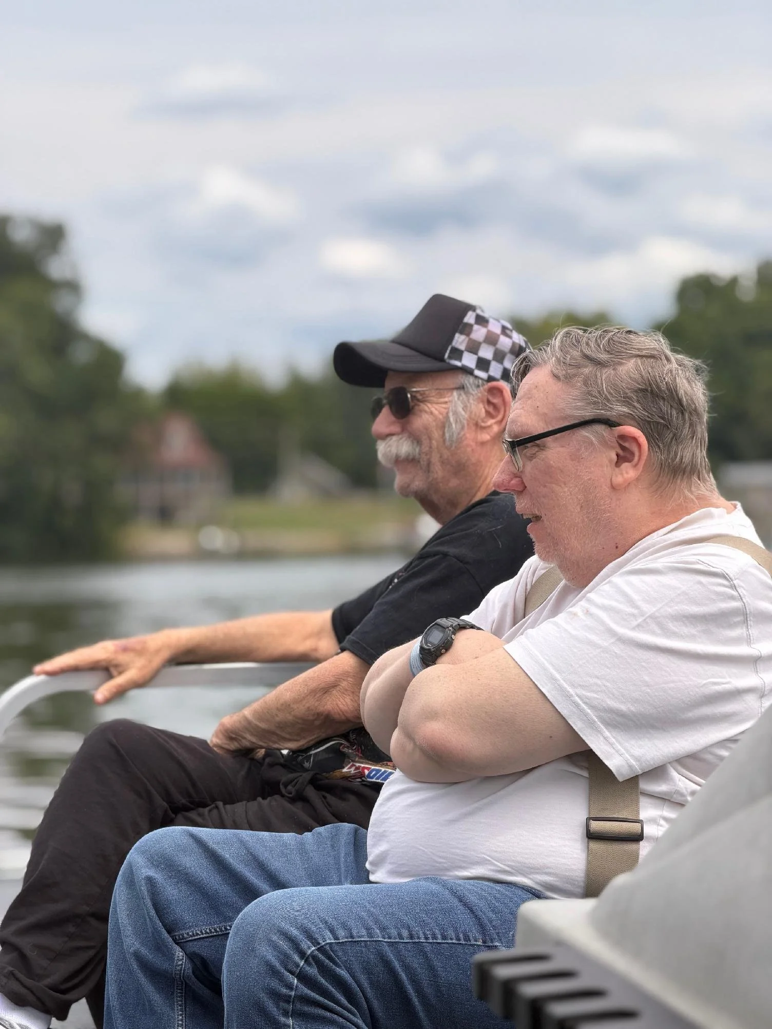 Two men sitting outdoors on a bench near water, with trees and cloudy sky in the background. One man is wearing sunglasses and a baseball cap with a checkered pattern on the side, the other has glasses and watch, both appear relaxed.
