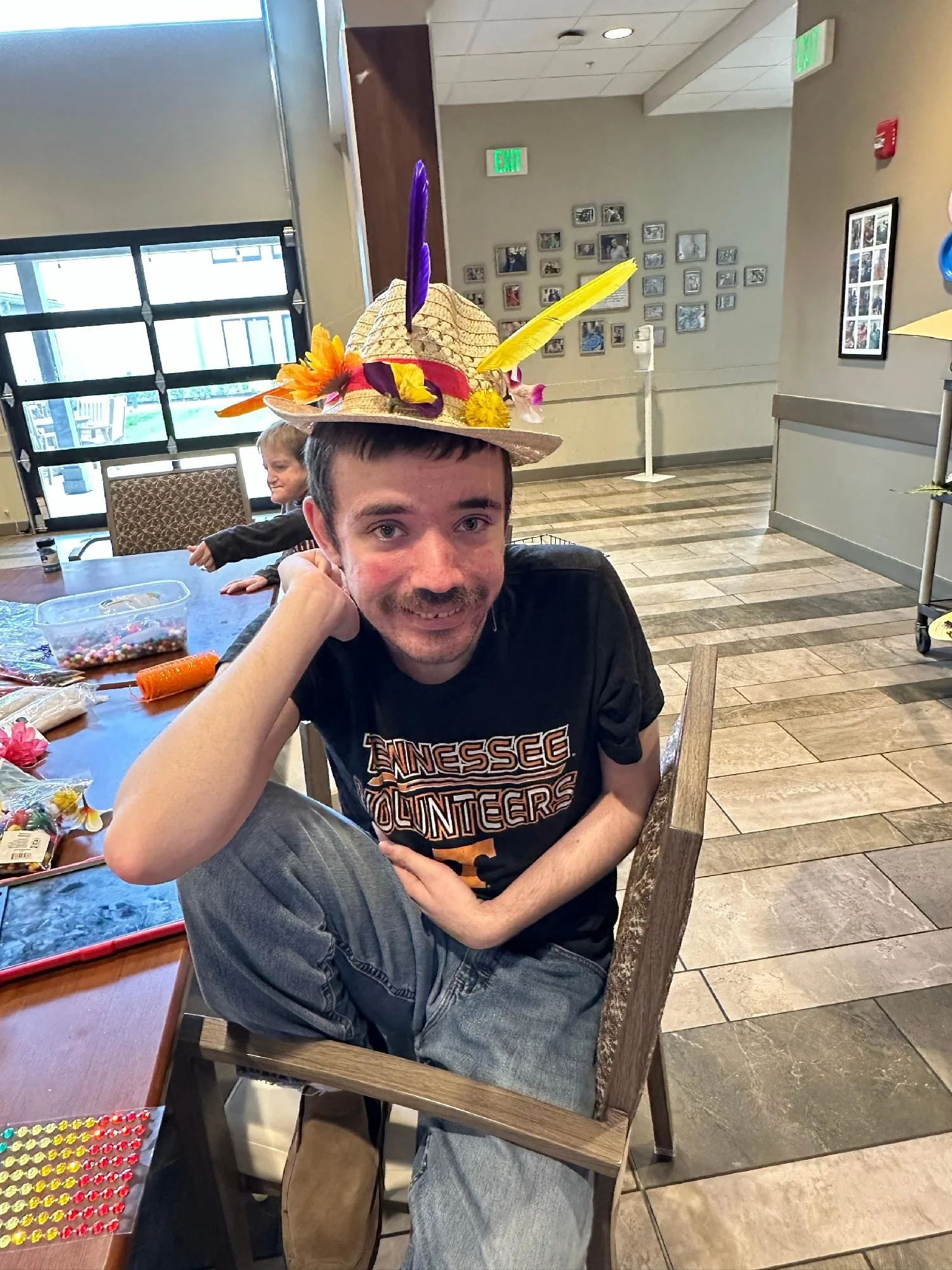 A young man sitting at a table wearing a large, colorful, decorated hat with feathers. The table has craft supplies such as beads and yarn. A child is in the background, sitting at the same table.