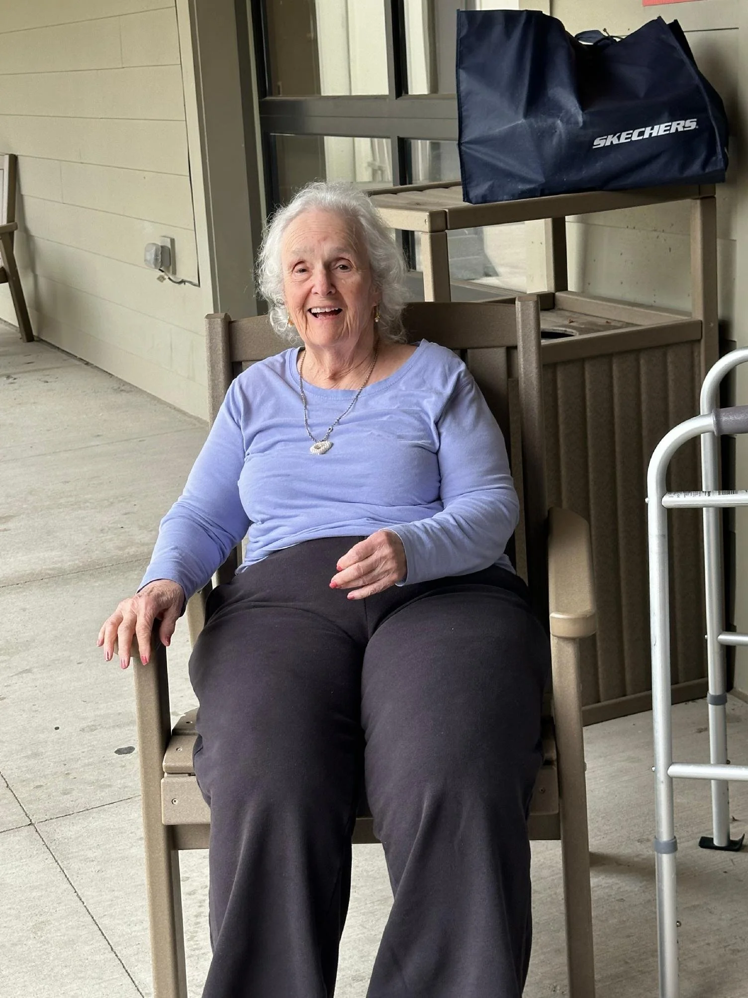 An elderly woman with white hair, sitting in a beige wooden chair on a porch, smiling and looking at the camera. There is a blue Skechers bag on a small shelf behind her, and a walker nearby.
