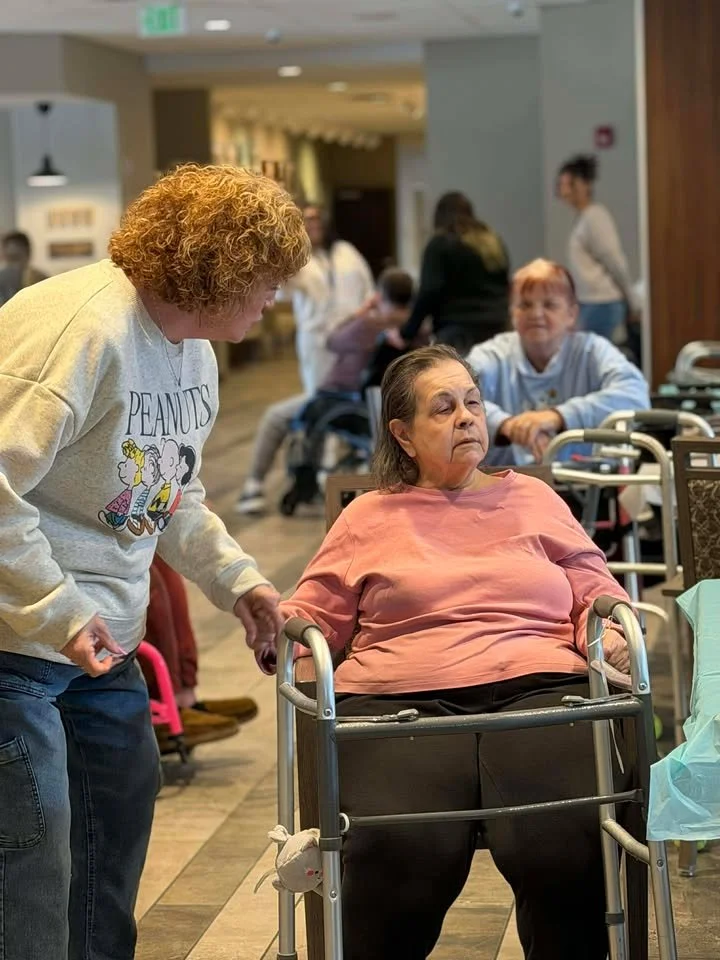 A caregiver interacts with an elderly woman in a wheelchair in a communal area of a nursing home or assisted living facility, with other residents and staff visible in the background.
