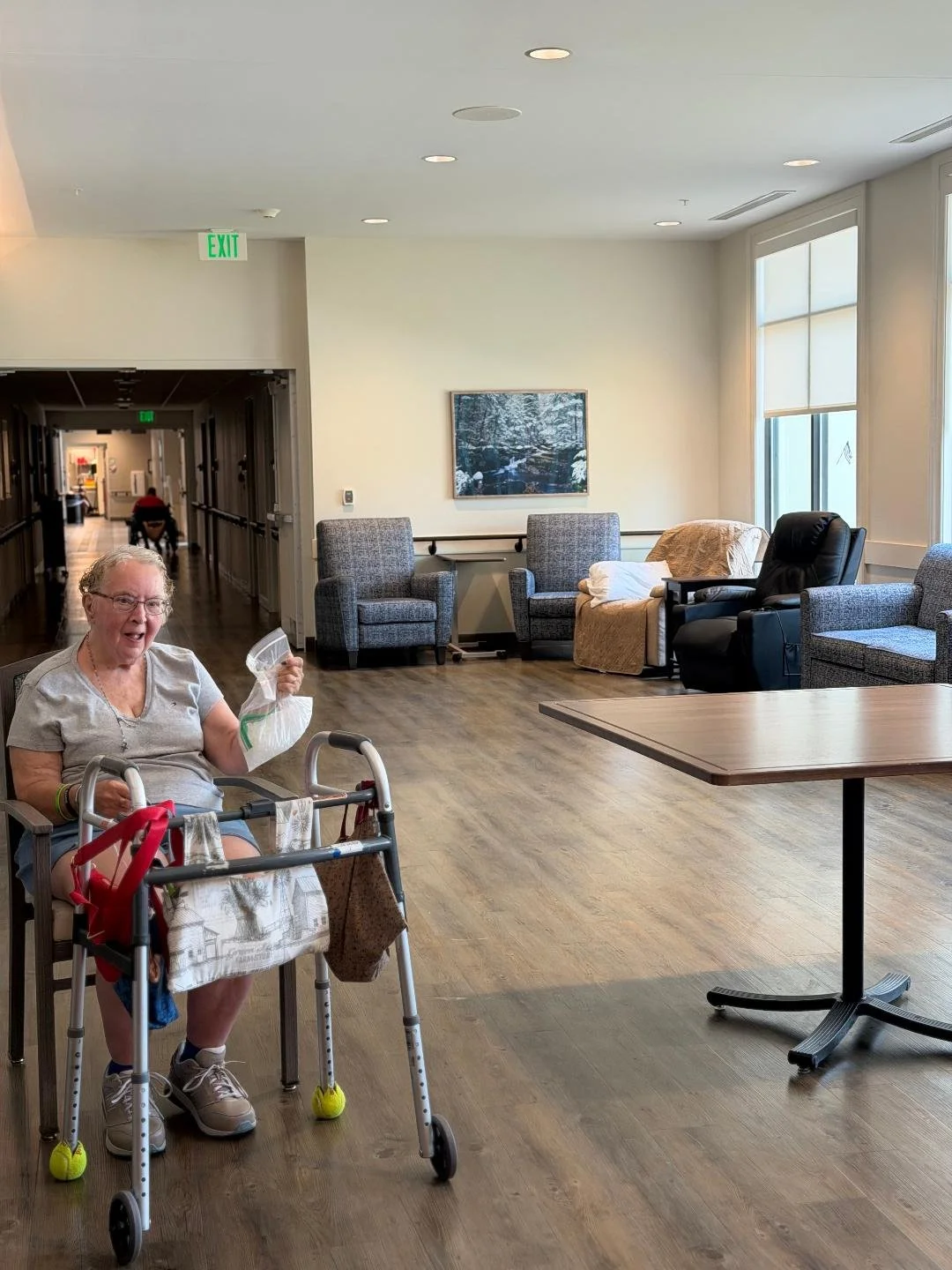 An elderly woman with glasses, sitting in a chair with a walker, smiling and holding a plastic bag in a bright, spacious lounge area with multiple armchairs, large windows, and a framed painting of a snowy forest landscape.