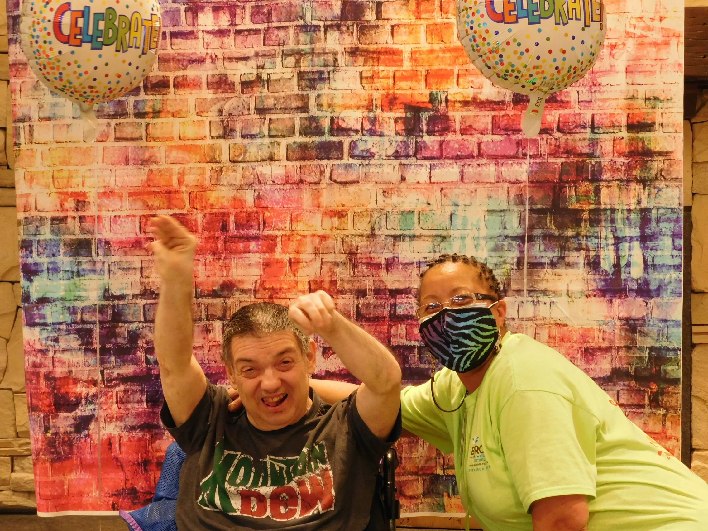Three people are celebrating in front of a brick-patterned background with balloons that say 'Celebrate.' Two people are in the foreground, one smiling with a black shirt, and the other wearing a bright yellow shirt and a mask. The third person's face is partially visible at the bottom of the photo.