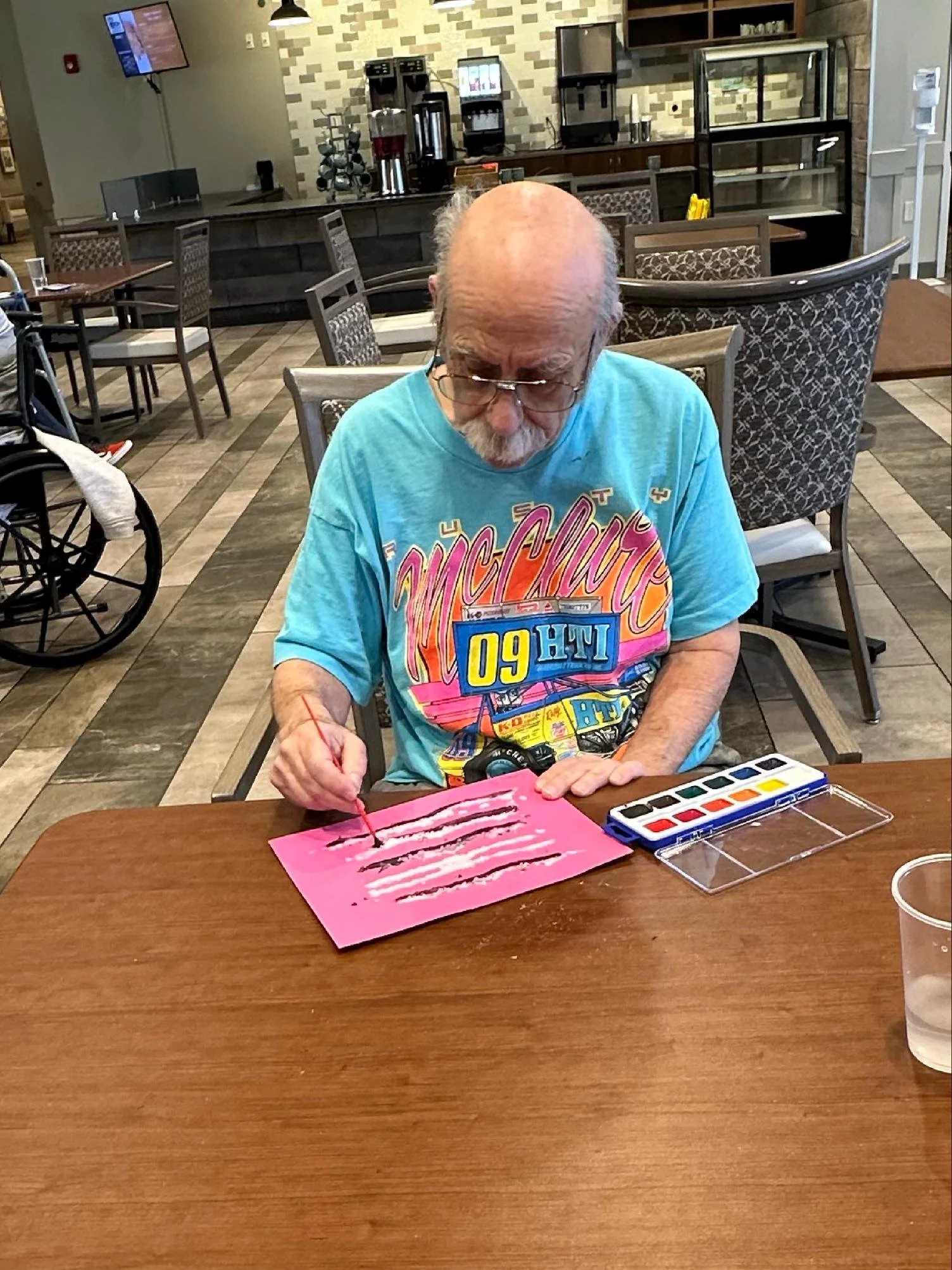 An elderly man with glasses and a beard, wearing a colorful graphic t-shirt, sits at a table in a restaurant or cafe, painting or drawing on a pink piece of paper with watercolor paints nearby. The background features a counter area with coffee machi