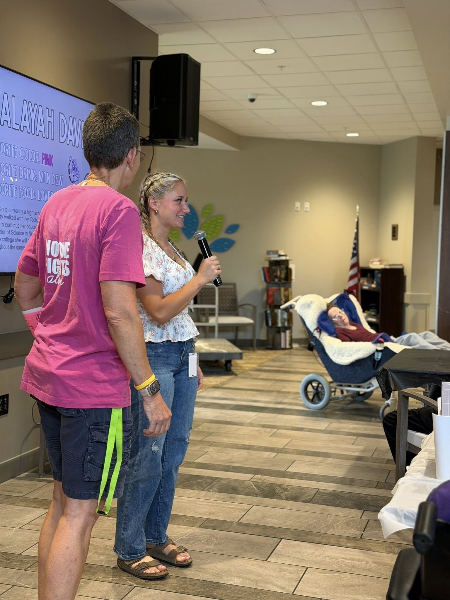 Three people standing in a room with one woman speaking into a microphone, two others looking at her, an elderly person resting in a wheelchair, and a large screen display in the background.
