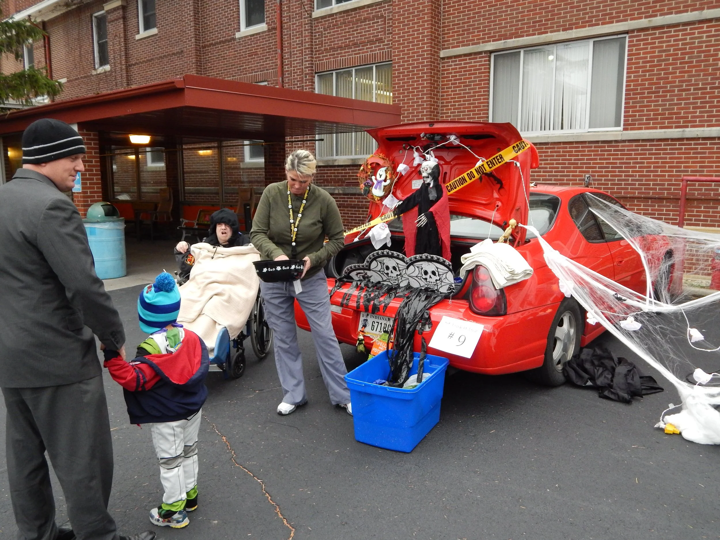 People in Halloween costumes standing near a decorated red car with spooky decorations, police tape, and cobwebs, in front of a brick building