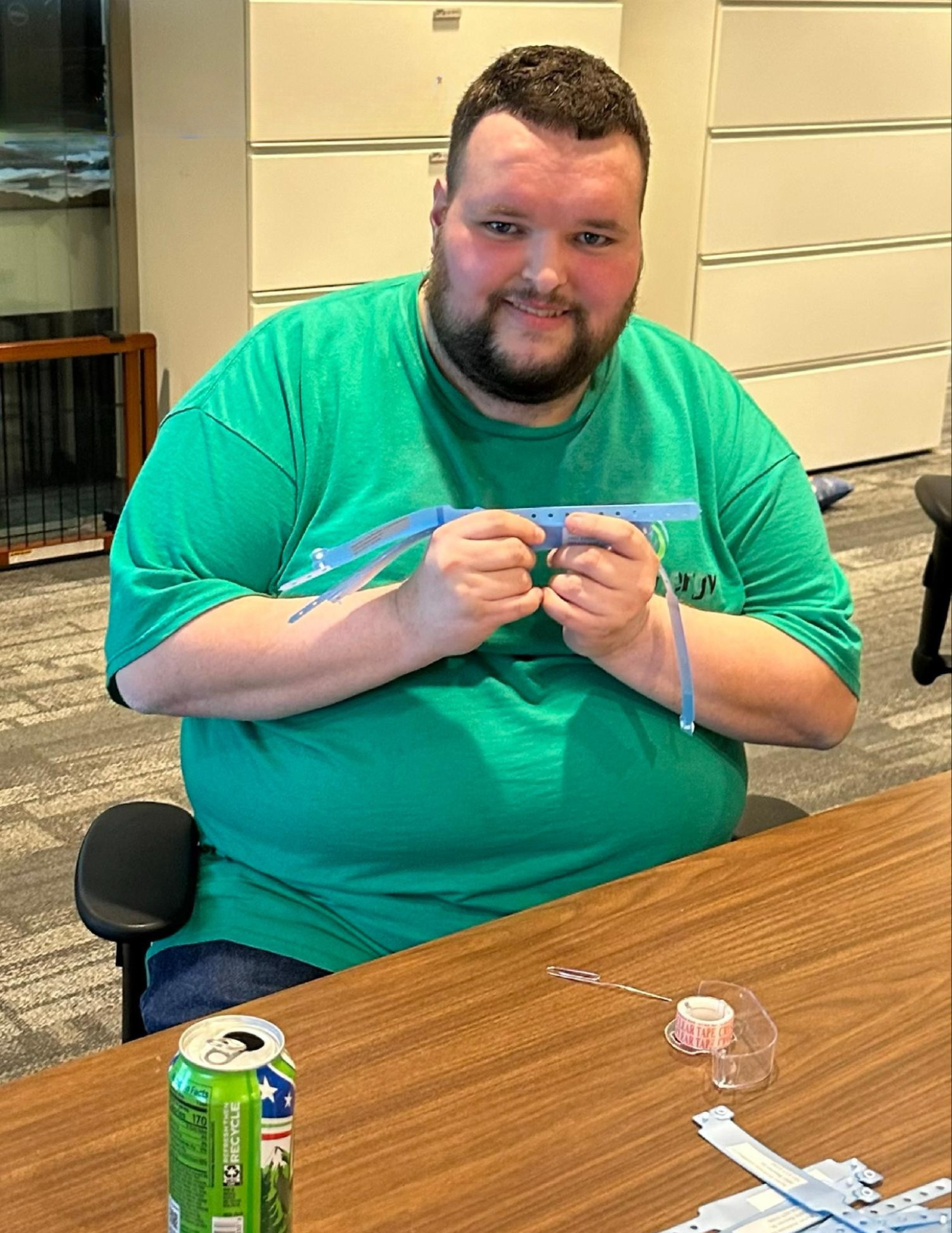 A man with a beard and short dark hair, wearing a green t-shirt, is sitting at a wooden table. A can of Sprite soda and some electrical components are on the table in front of him. He is holding a piece of plastic or a strap. The background shows a m