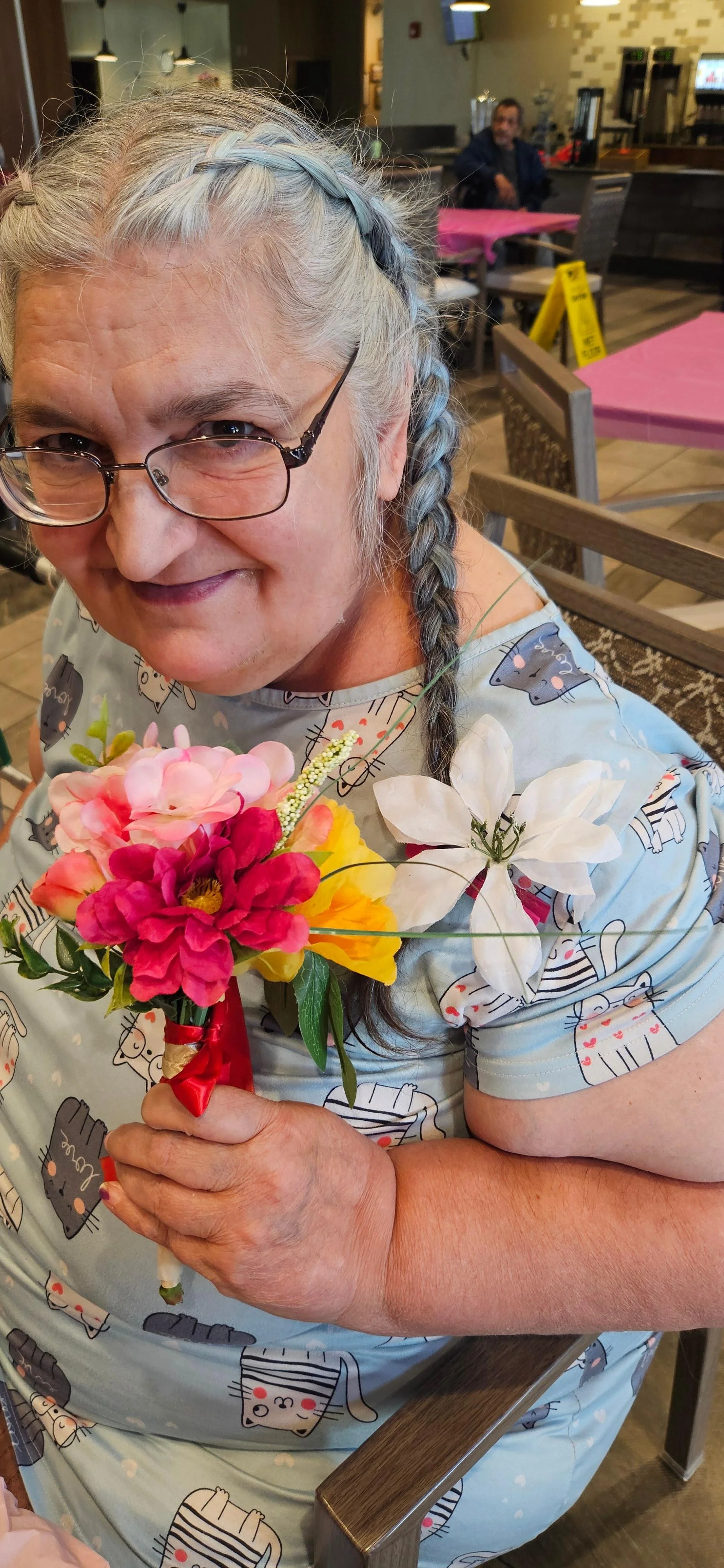 An elderly woman with gray hair in a braid, wearing glasses, holding a bouquet of colorful flowers, sitting in a restaurant or cafe, smiling at the camera.