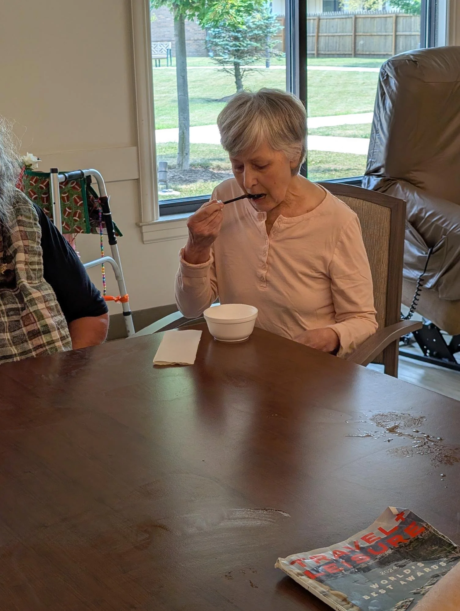 An elderly woman with short gray hair sitting at a wooden table near a window, eating with a spoon from a white bowl. There is a magazine or booklet on the table and a walker with a patterned cloth in the background. Outside the window, a green yard 