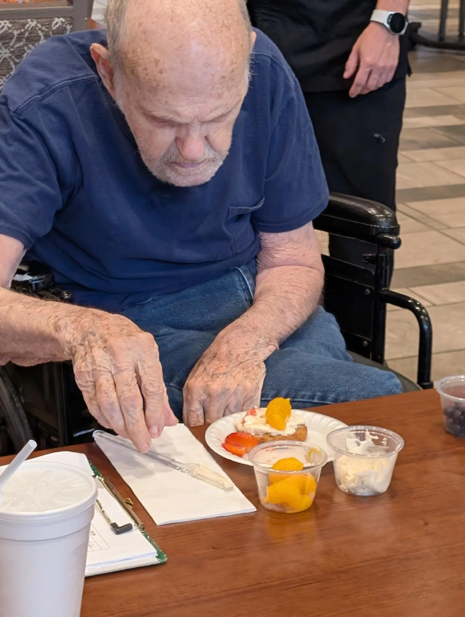 An elderly man with white hair and a beard, wearing a blue t-shirt, sitting at a wooden table. He is preparing a dessert with strawberries, whipped cream, and a yellow fruit on a paper plate. Several small plastic containers with fruit and whipped cr