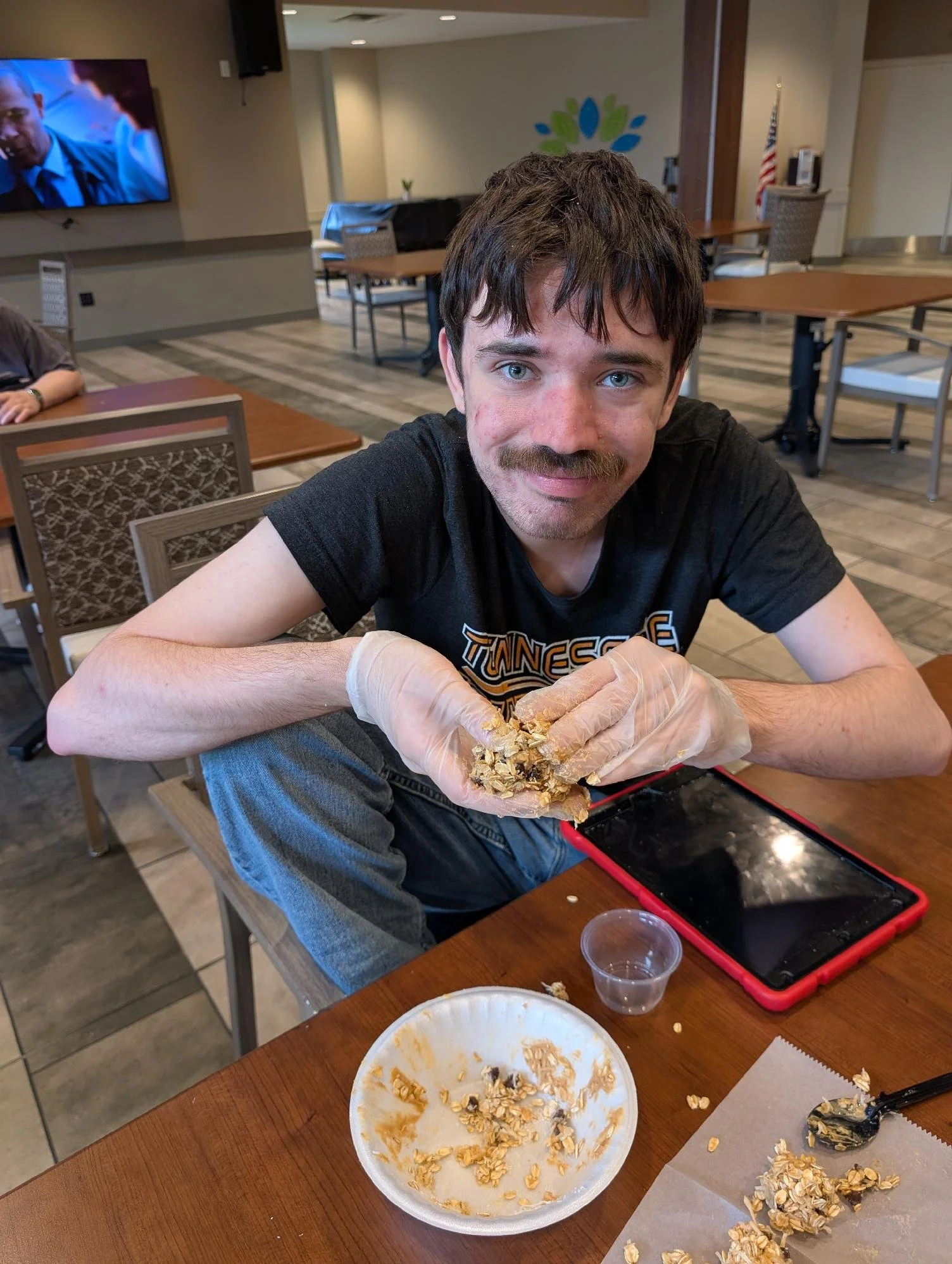 A young man with blue eyes and a mustache, wearing a black T-shirt, is sitting at a wooden table holding a handful of oats or granola. There is a white disposable plate with oats on it in front of him, along with some scattered oats and a black spoon