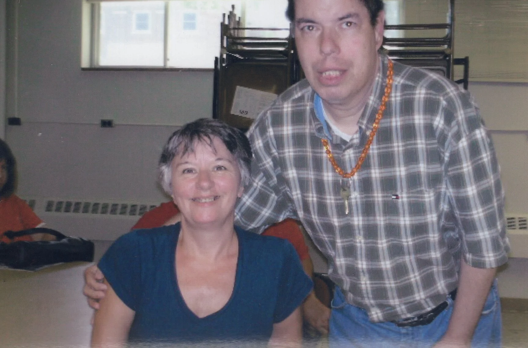 A woman with short gray hair smiling and a man with dark hair standing beside her, both indoors in a room with chairs and a window in the background.