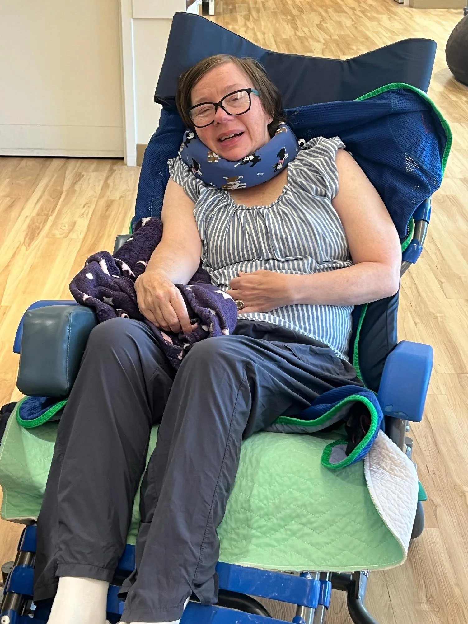 A woman with glasses sitting in a wheelchair, smiling and wearing a striped shirt and a floral neck scarf. She has a blanket over her lap, and there is a green blanket underneath her feet.