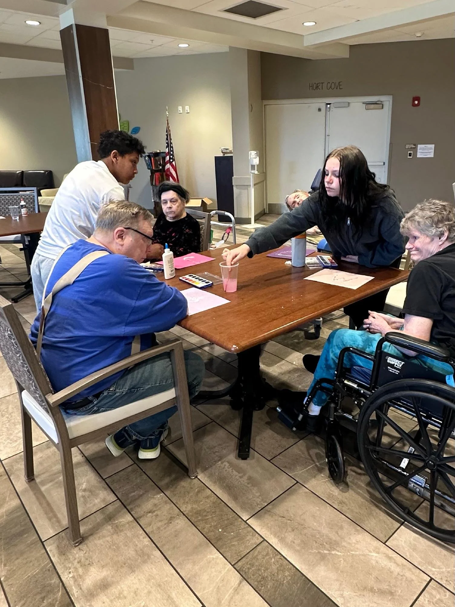 A group of people, including a young man, an elderly woman, a middle-aged man in a wheelchair, and two women, are gathered around a wooden table in a room labeled 'Hort Cove'. The table has various items, including papers, a calculator, and a drink. 