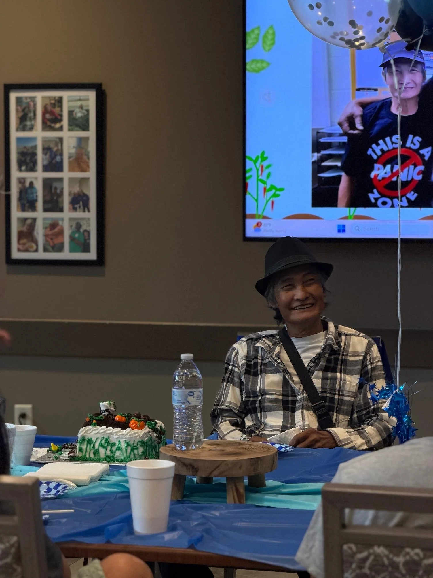 A smiling elderly woman wearing a black hat and plaid shirt seated at a celebration table with cake, balloons, and party decorations. A large cake with green and orange decorations is in front of her, and a water bottle is visible on the table. A fra