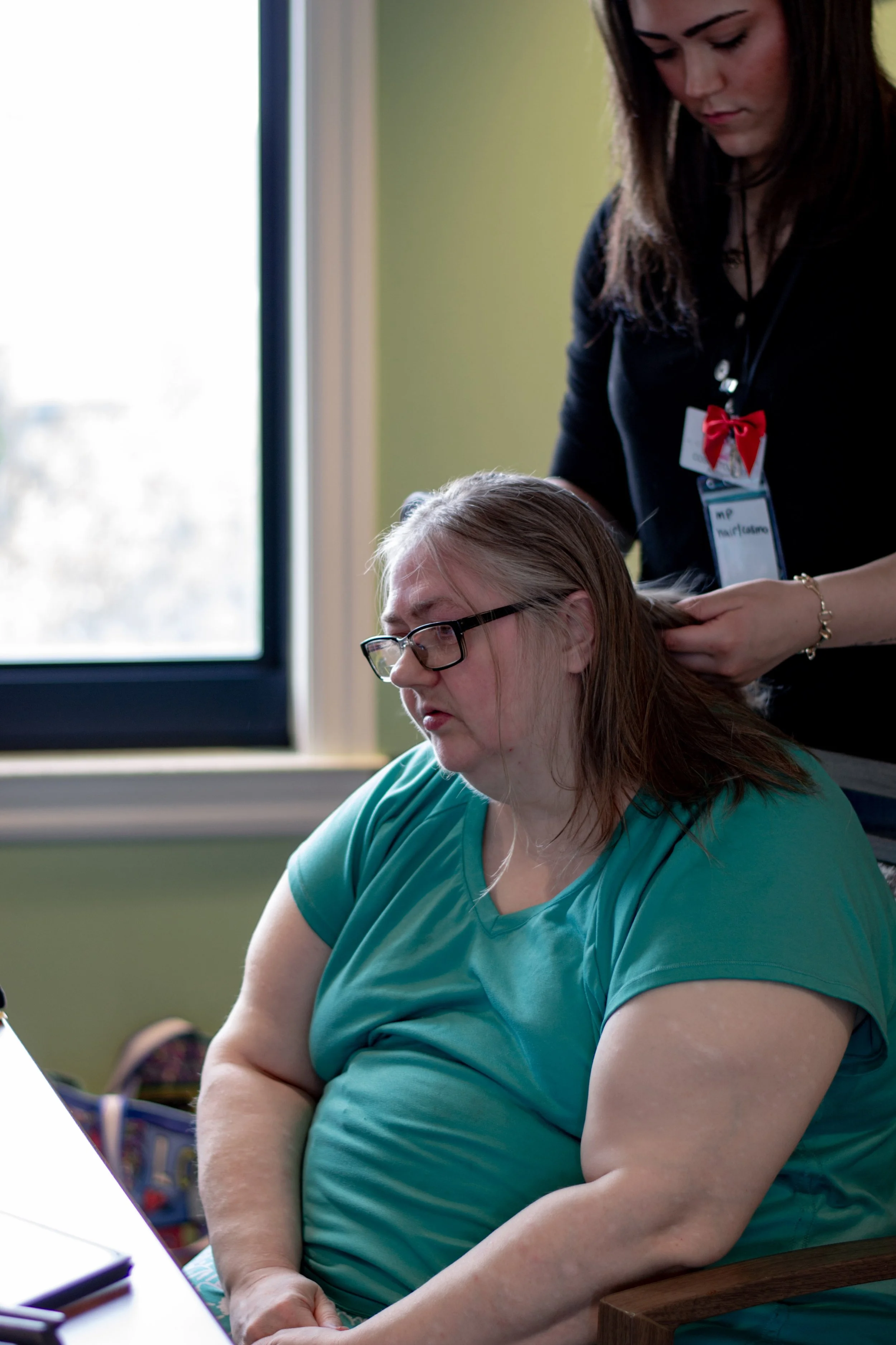 A middle-aged woman with glasses and green shirt sitting at a table with her arms crossed, looking down. A woman with dark hair, identifying badge with a red ribbon, is standing behind her, gently holding her shoulder.