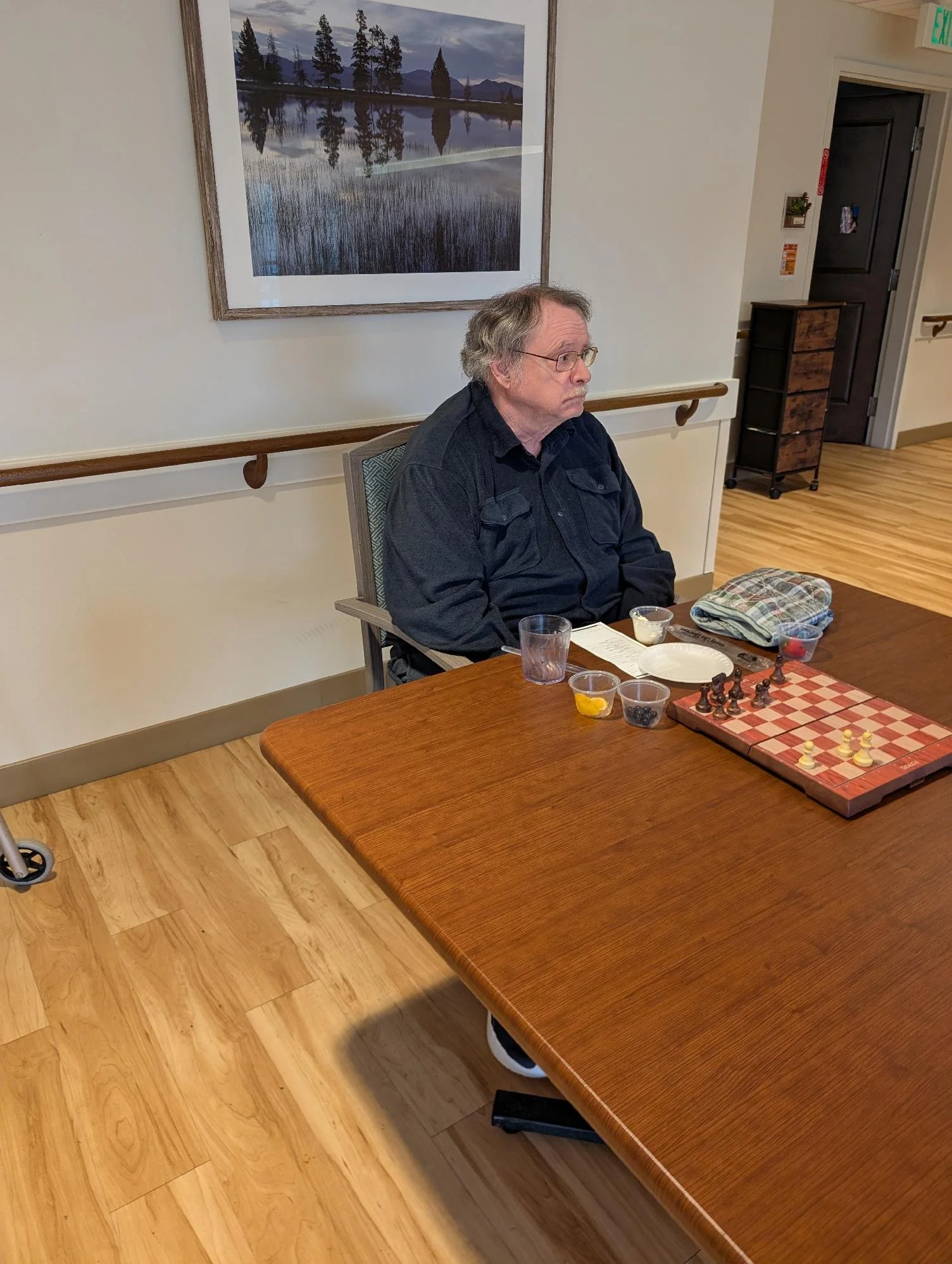An older man with gray hair, glasses, and a mustache, sitting at a wooden table with a chess game, snacks, and drinks. The room has light-colored walls, a large framed landscape photo of trees and water, and a dark door in the background.