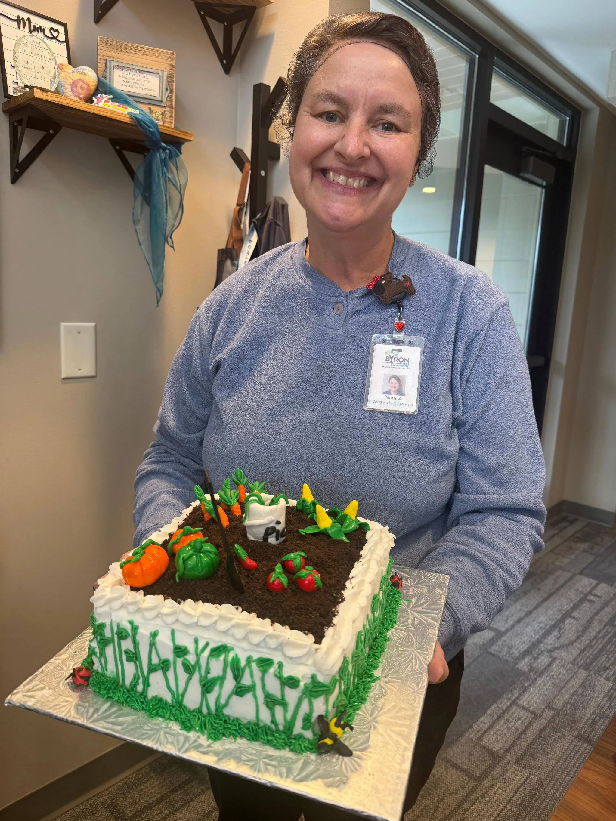 A smiling woman holding a decorated cake with a gardening theme, featuring small pumpkin and tomato frosting decorations and green piped grass icing around the base, inside a building near a window.