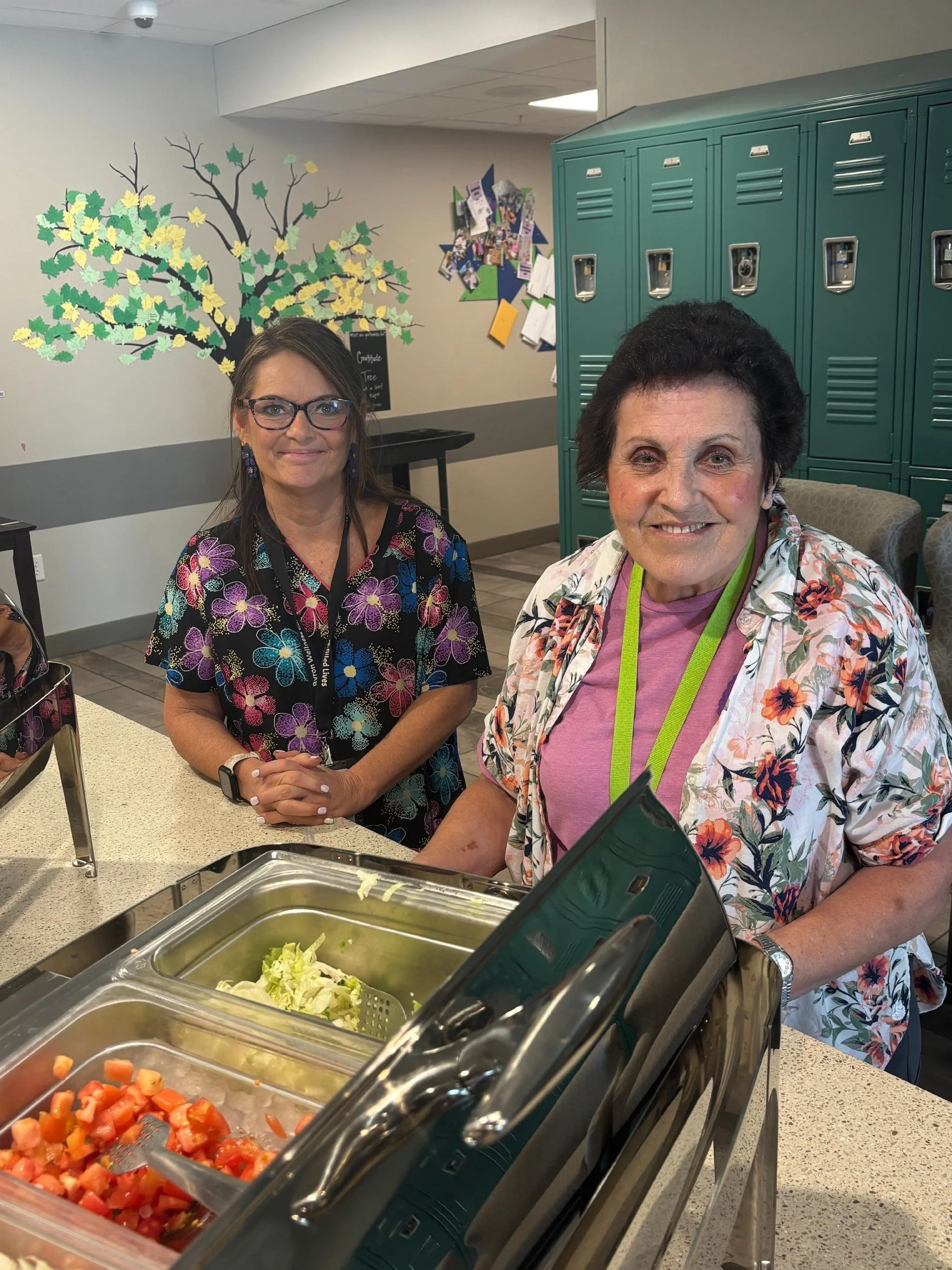 Two women smiling at a cafeteria buffet with trays of chopped vegetables, including tomatoes and lettuce. One woman has glasses and a floral shirt, the other has short dark hair and a floral jacket. There are teal lockers and colorful bulletin boards