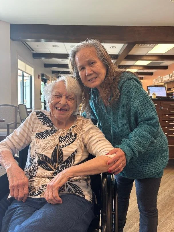 A young woman is holding the hand of an elderly woman in a wheelchair, both smiling in a well-lit indoor setting.