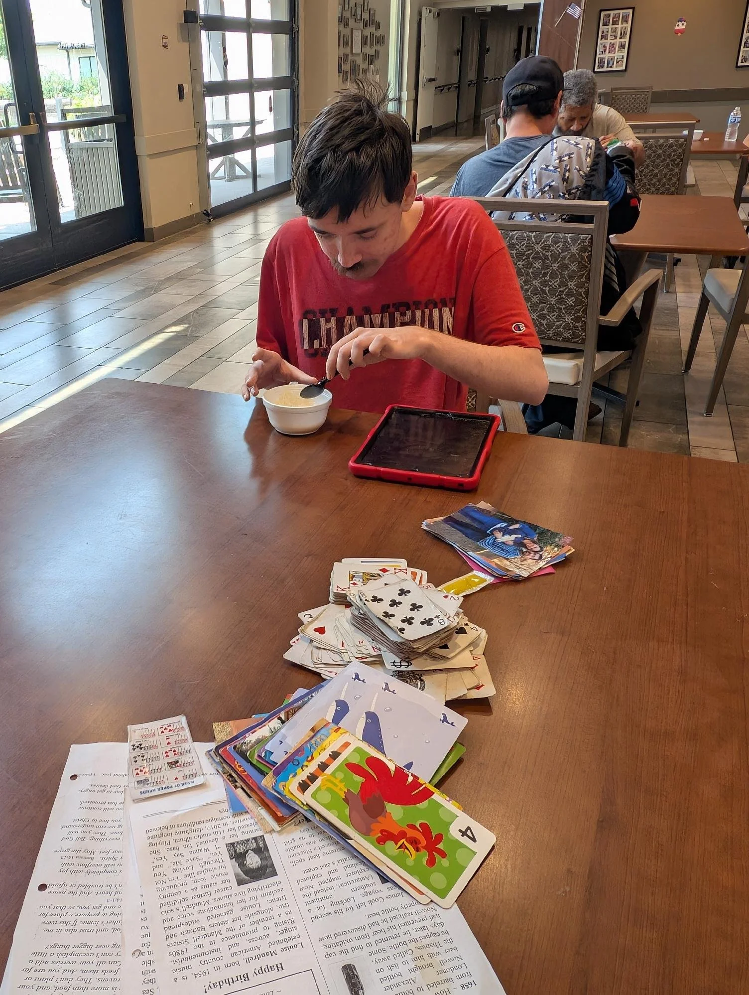 A person in a red shirt sitting at a wooden table, looking down at a bowl and a tablet. There are scattered coloring books, papers, and playing cards on the table. In the background, other people are seated, and a glass door or window is visible.