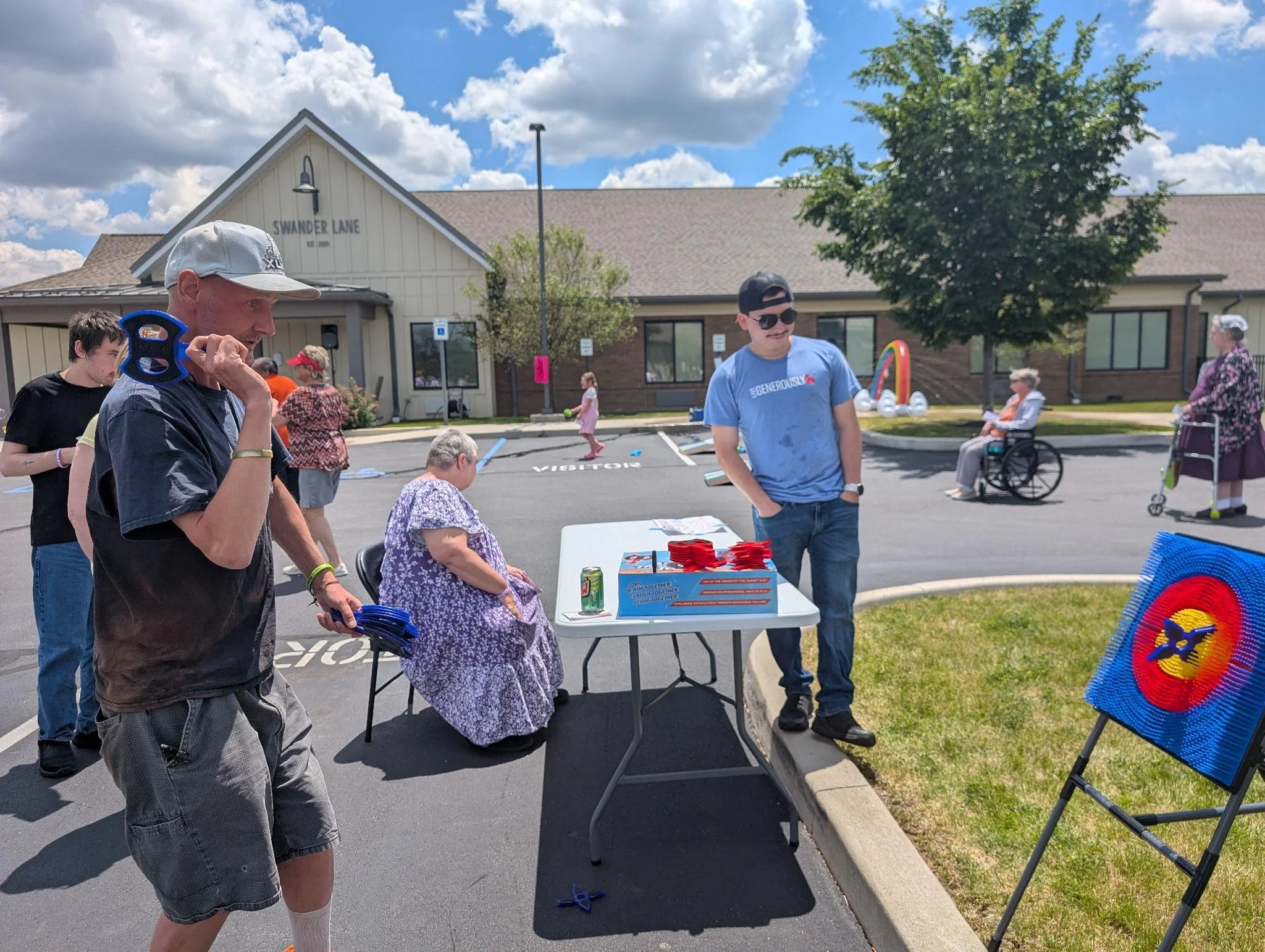 People participating in an outdoor event on a sunny day in a parking lot outside a building named 'Swander Lane.' Some are standing, sitting at tables, and managing booth activities, while others are walking or sitting, with a decorative rainbow arch and colorful items on display.
