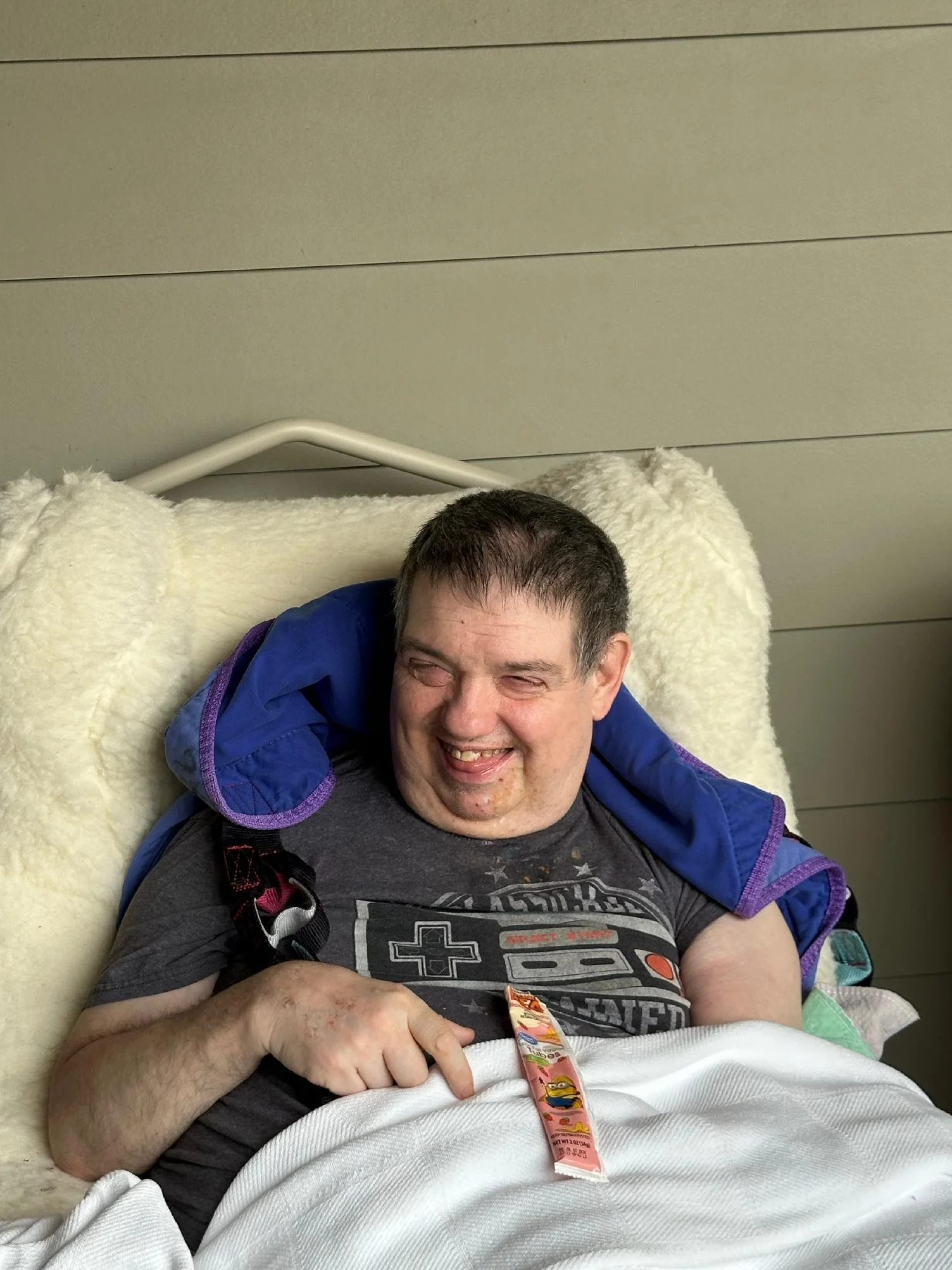 A smiling man lying in a hospital bed with a medical band on his wrist, a black t-shirt, a gray shirt with a graphic print, a blue jacket, and a blanket covering him.