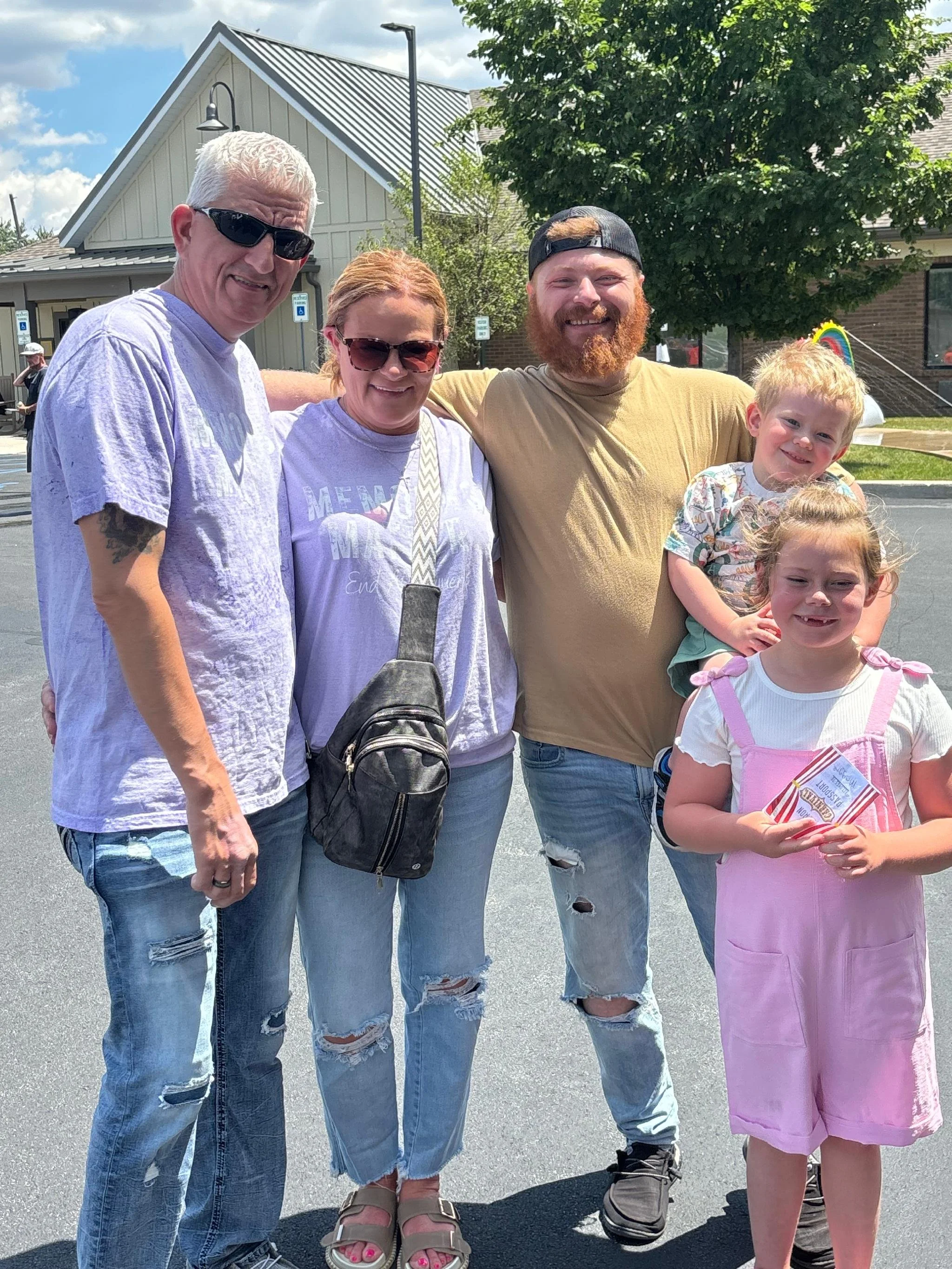 Group of four people standing outside on a sunny day, including three adults and two children, smiling at the camera, with a building and a rainbow-shaped decoration in the background.