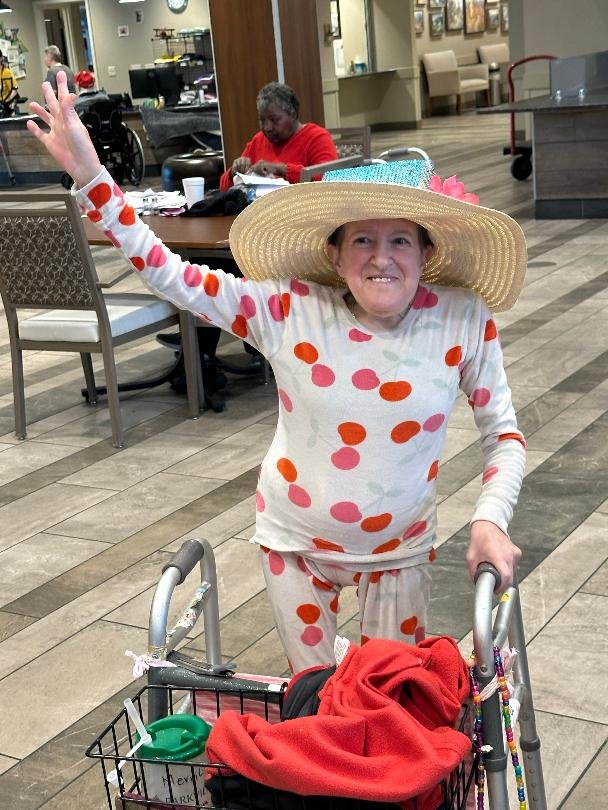 An elderly woman wearing a large straw sunhat and matching polka dot pajamas, standing with a walker in what appears to be a common area of a care facility or hospital, smiling and waving.