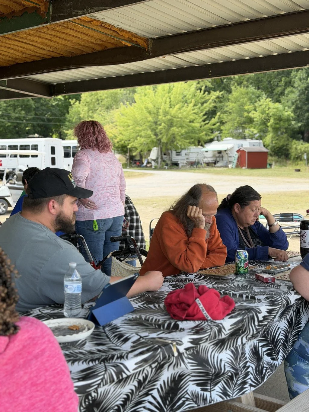 People sitting at a table outdoors with trees and trailers in the background, under a roofed shelter.