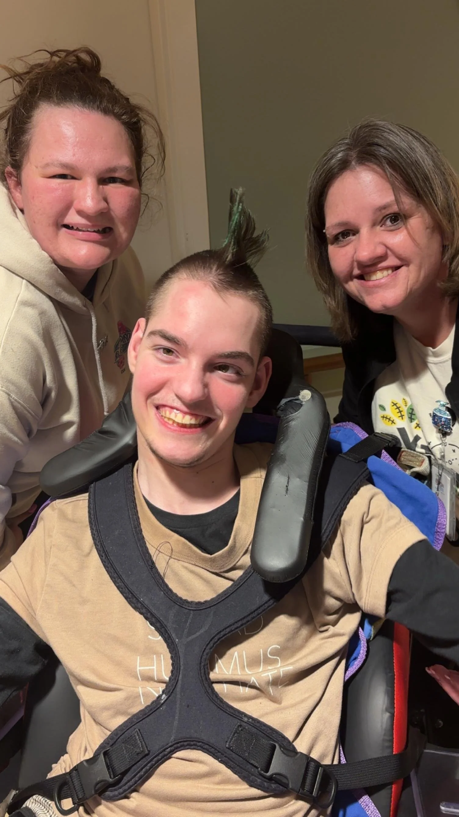 Three smiling people, one in wheelchair with a neck brace, two women standing behind him, one with messy hair, inside a room.