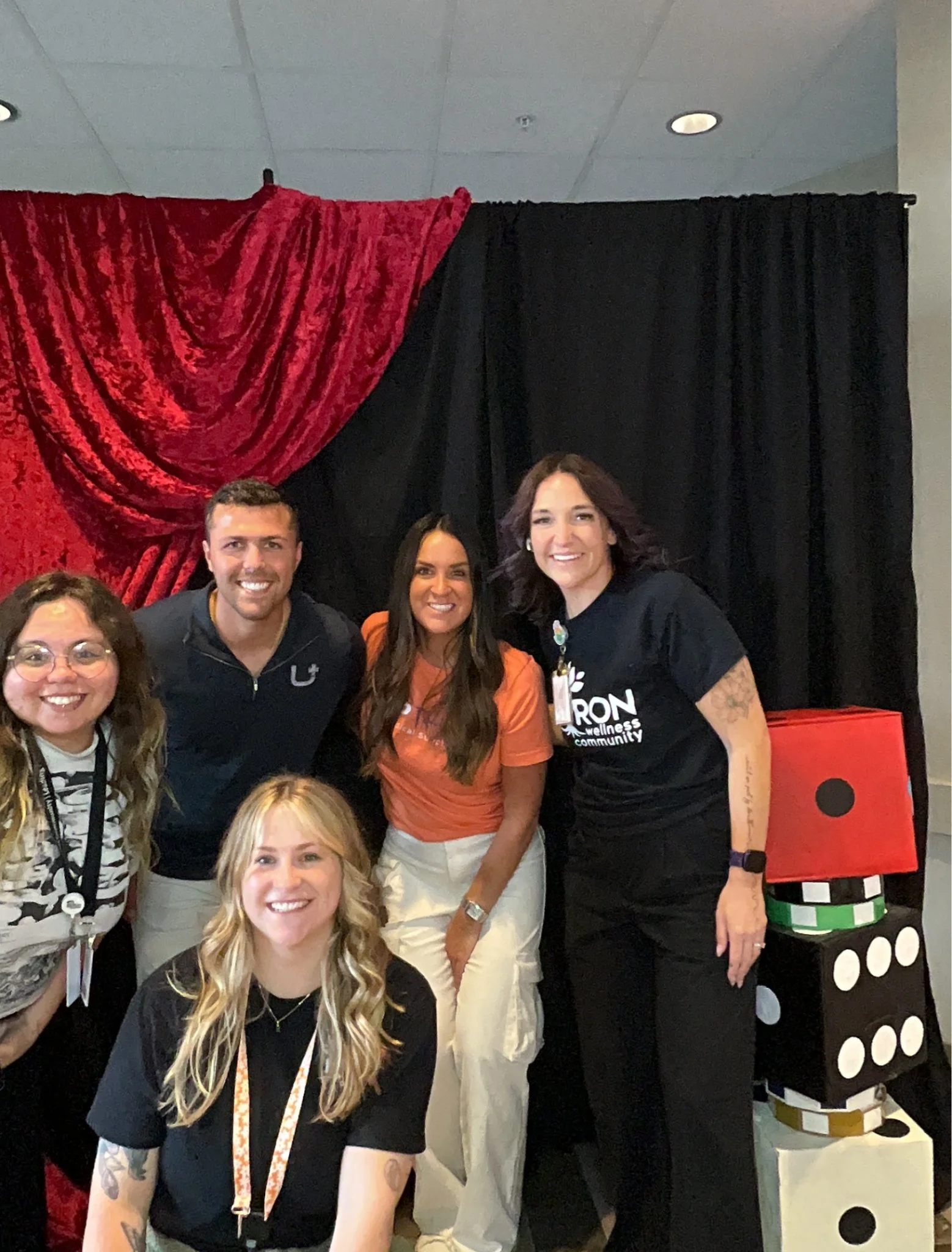 Group of people, mostly women, smiling and posing for a photo in front of a black and red curtain backdrop, with some dressed in casual attire and others in event or work-related clothing.