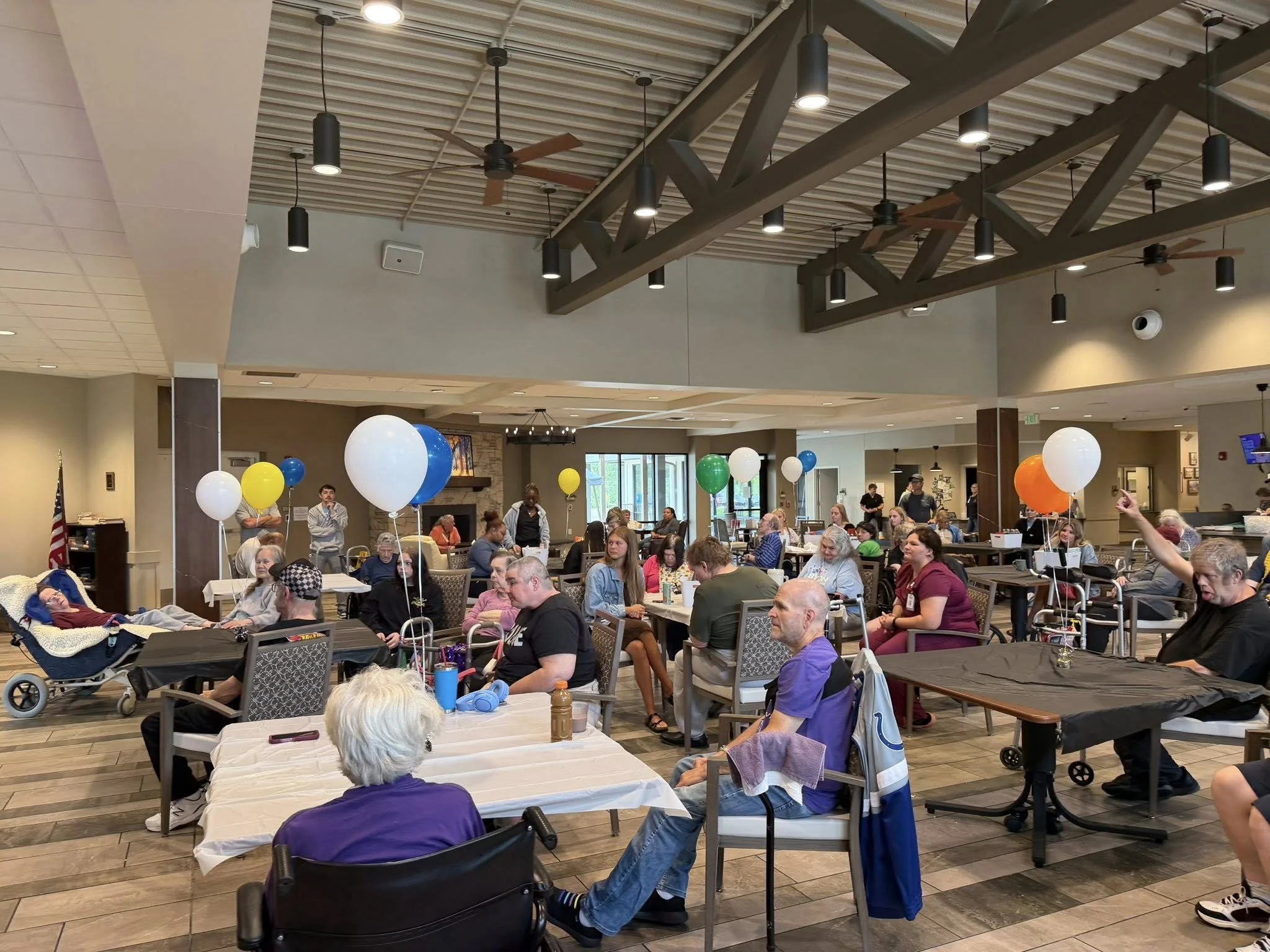 People sitting at tables in a spacious room with balloons, some people are in wheelchairs, and others are standing or walking, with a high ceiling, hanging lights, and a fireplace in the background.