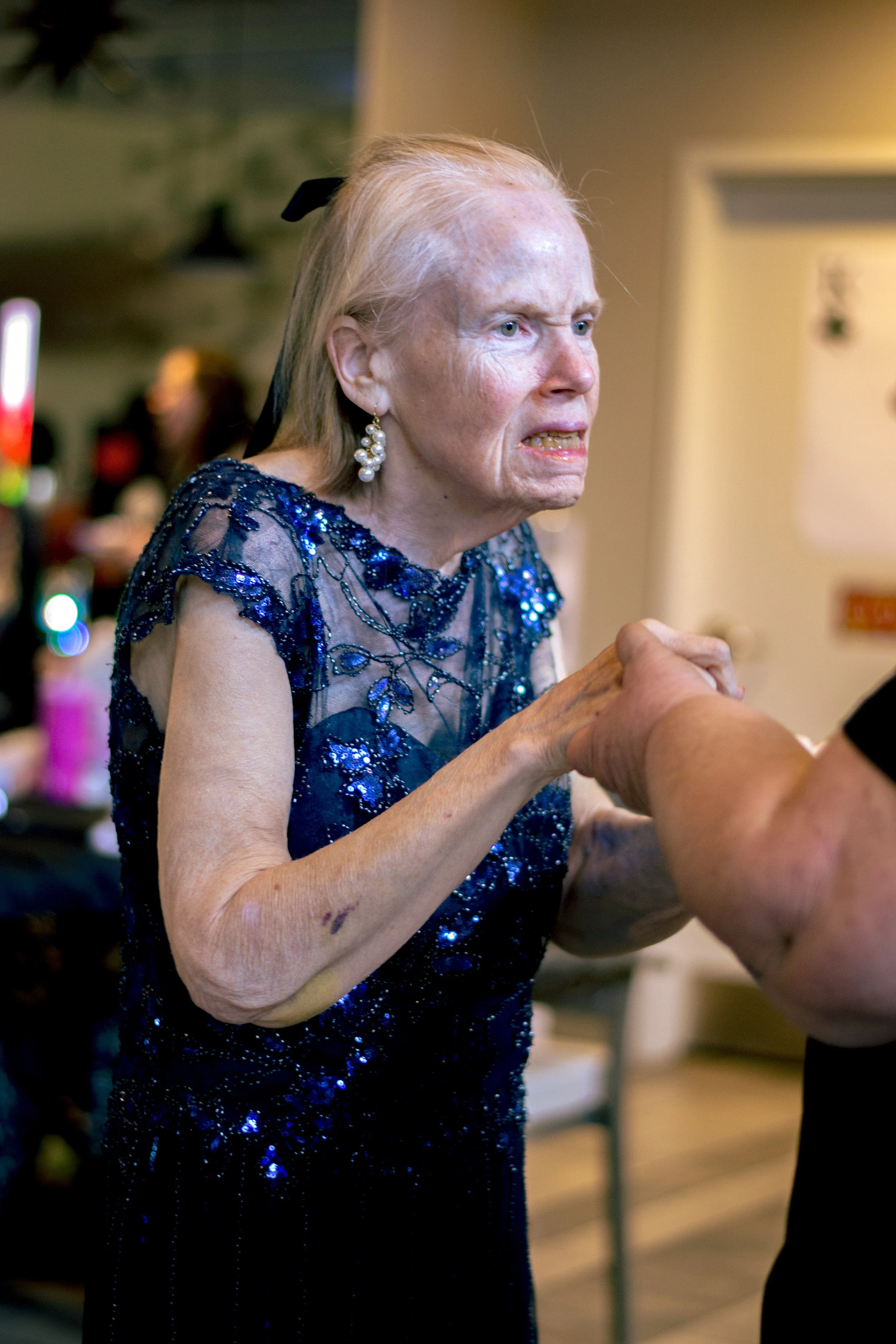 An elderly woman with white hair, wearing a blue sequined dress and pearl earrings, holding hands with someone outside the frame.
