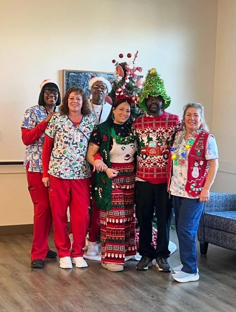 Group of six people celebrating Christmas, wearing festive holiday attire, standing in front of a decorated Christmas tree. Some are wearing Santa hats, Christmas sweaters, and holiday-themed accessories.