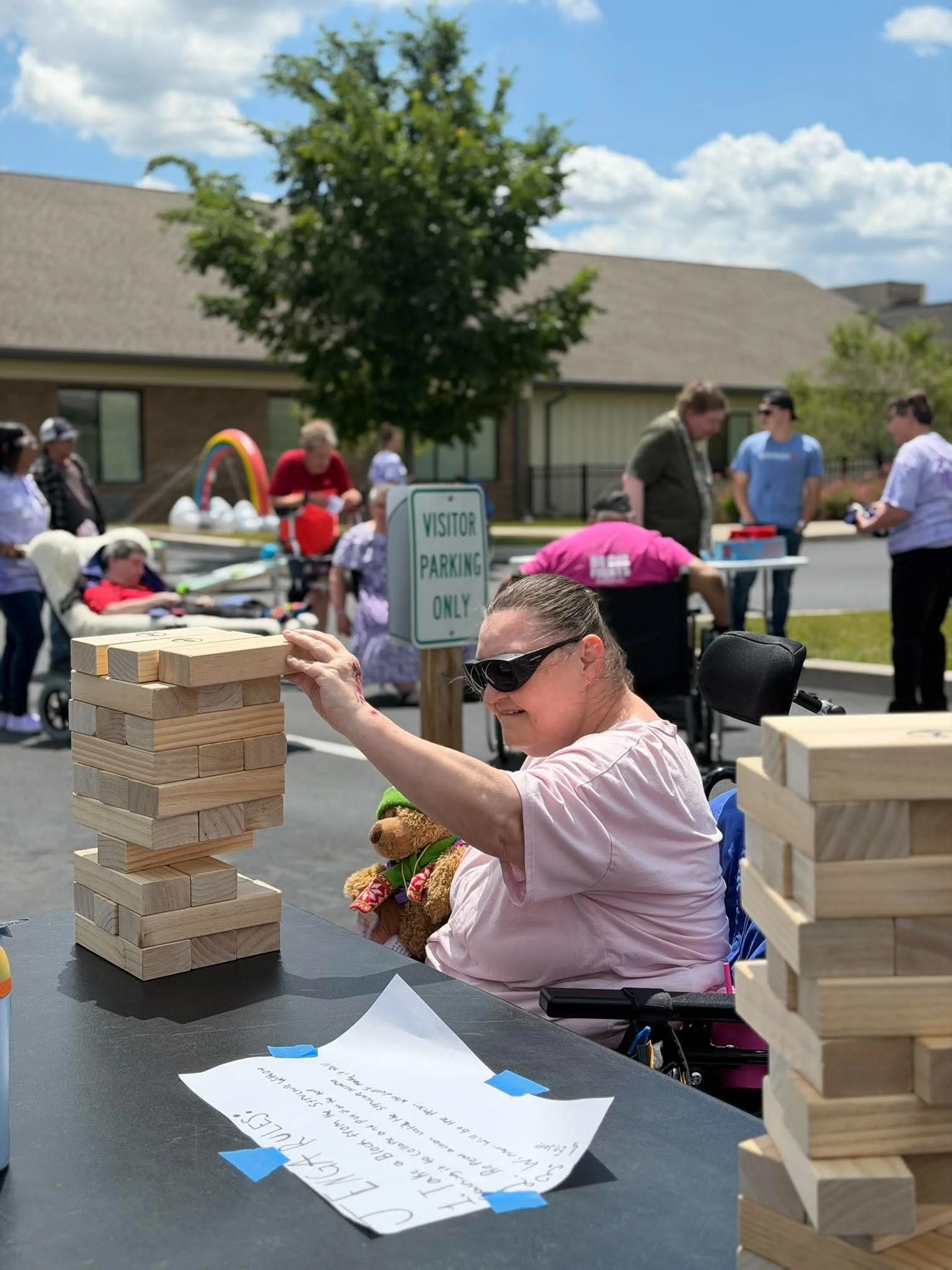 A woman in a pink shirt and sunglasses playing a large outdoor Jenga game, with a teddy bear on her lap. In the background, there are several people, a tree, a building, and a sign that reads 'Visitor Parking Only'. The sky is partly cloudy.