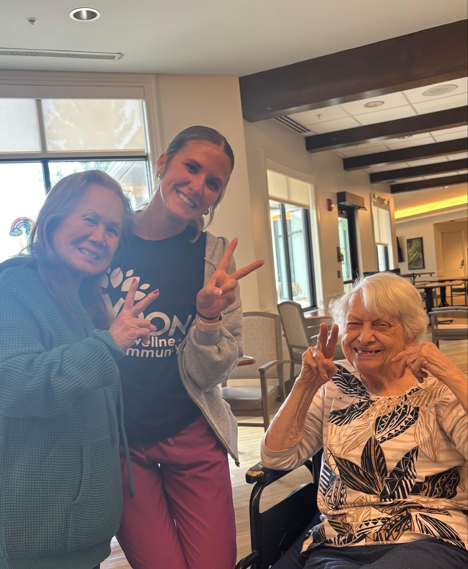 Three smiling women, two standing and one seated in a wheelchair, making peace signs in a well-lit indoor setting with large windows and wooden ceiling beams.