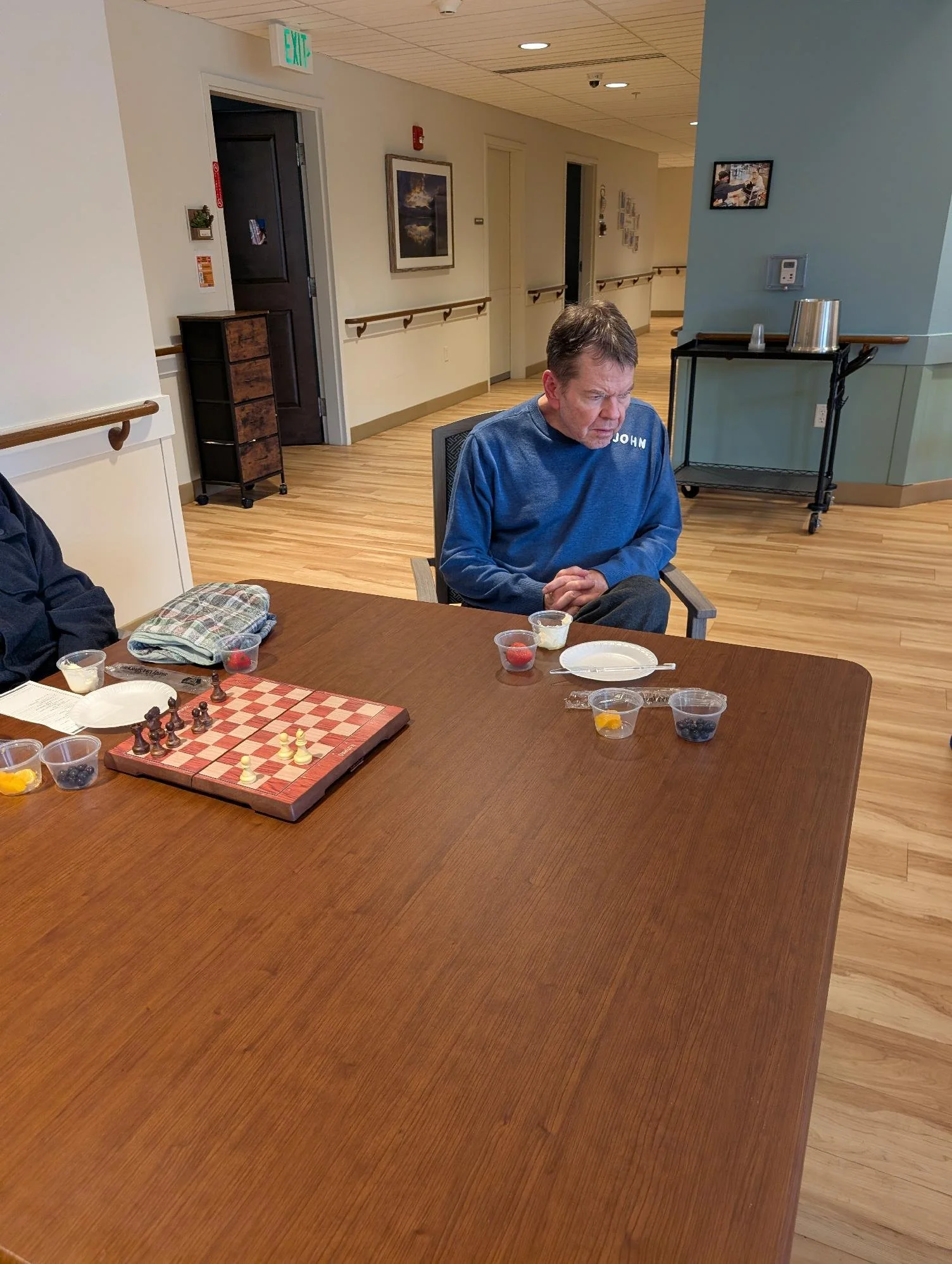 A middle-aged man with brown hair, wearing a blue shirt, sitting at a wooden table in a room, appearing focused on a chess game. The table has some plastic containers with strawberries and blueberries, and a checkered chessboard. The background shows