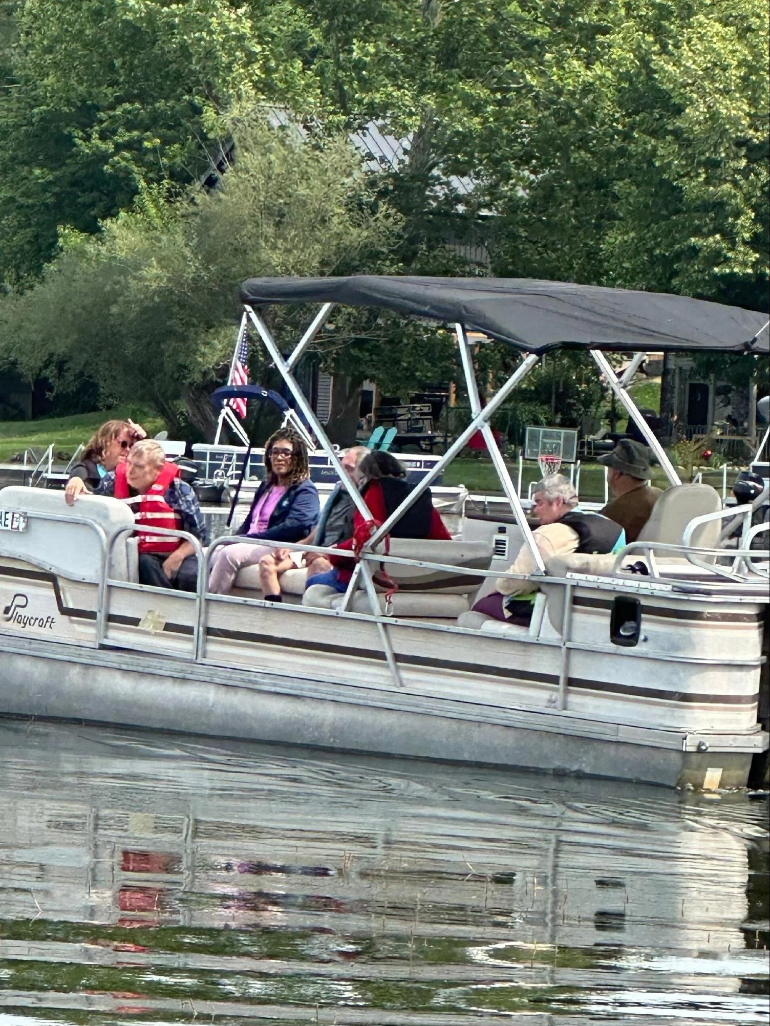 A boat with a canopy is on a body of water, carrying several people, some of whom are wearing life jackets. The boat appears to be moving along a river or lake, and there are trees and houses visible in the background.