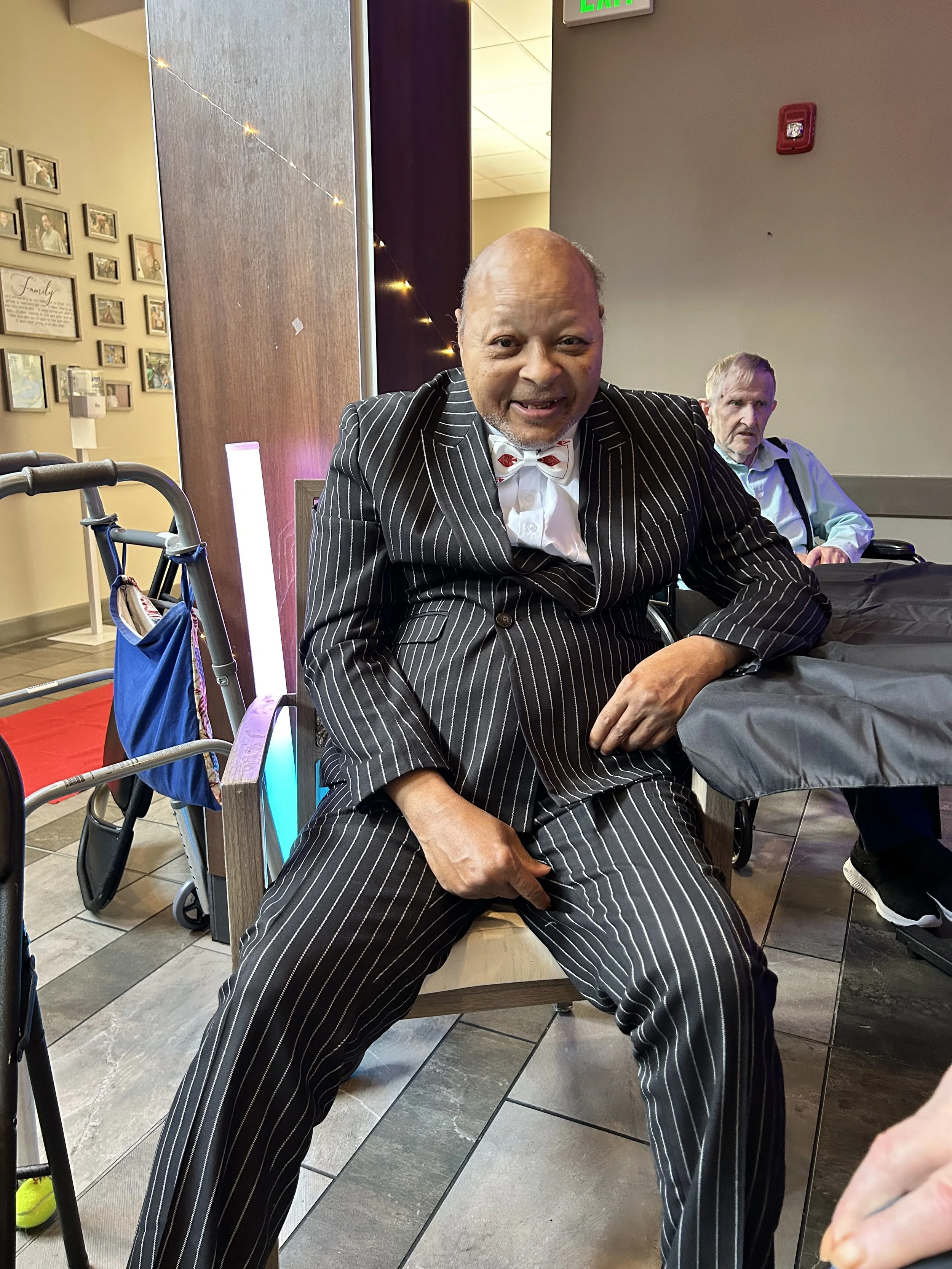 A man in a black pinstripe suit and bow tie sitting on a chair, smiling at the camera, with a woman in a wheelchair and a man in the background.