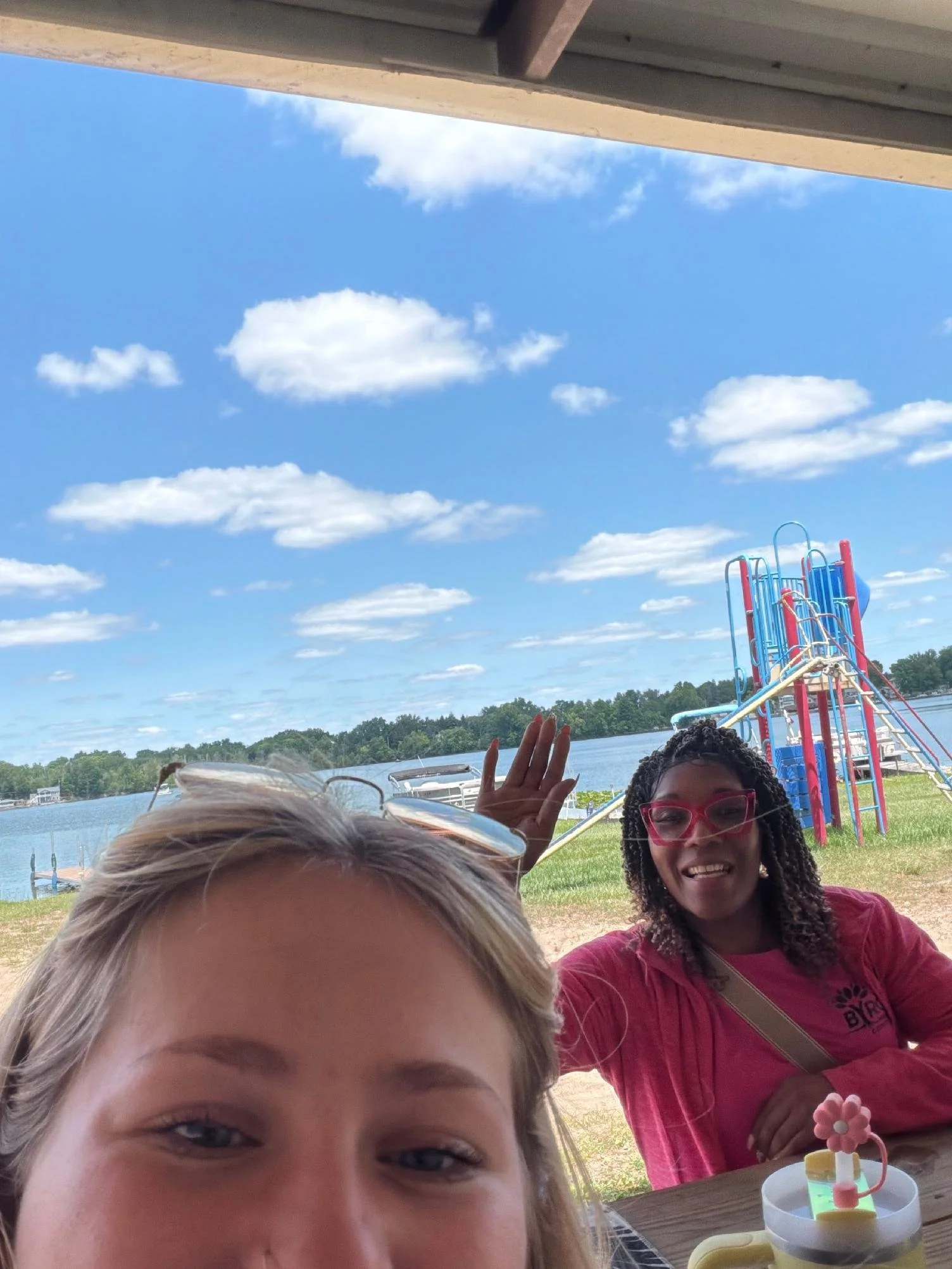 Partially visible young girl with blonde hair in the foreground, a smiling woman with dark hair and glasses in the background, blue sky with clouds, and a playground near a lake.