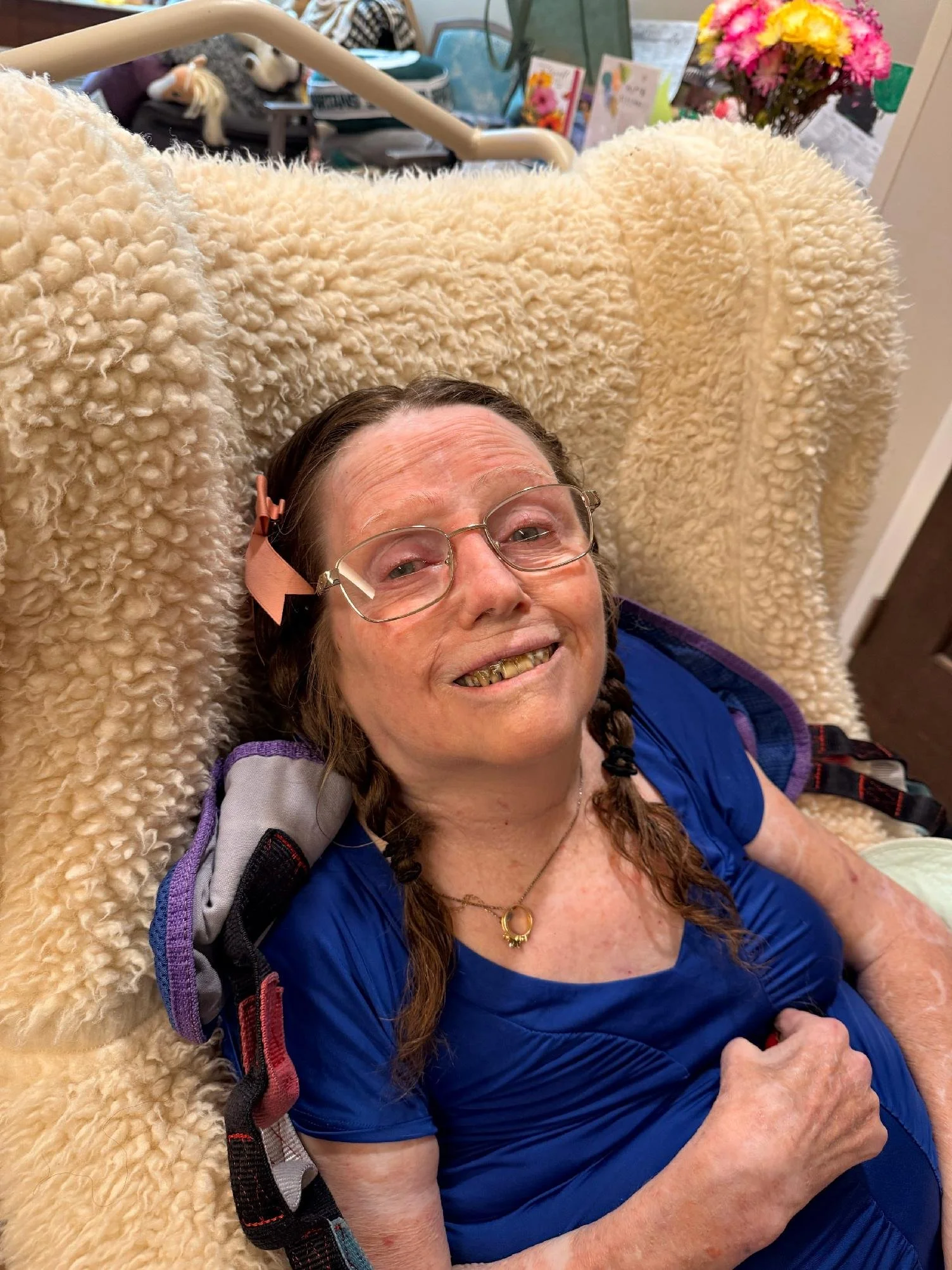 An elderly woman with glasses smiling, lying on a cream-colored fuzzy blanket or pillow, wearing a blue shirt, with a gift or harness strap around her shoulder, in an indoor setting with flowers and books in the background.