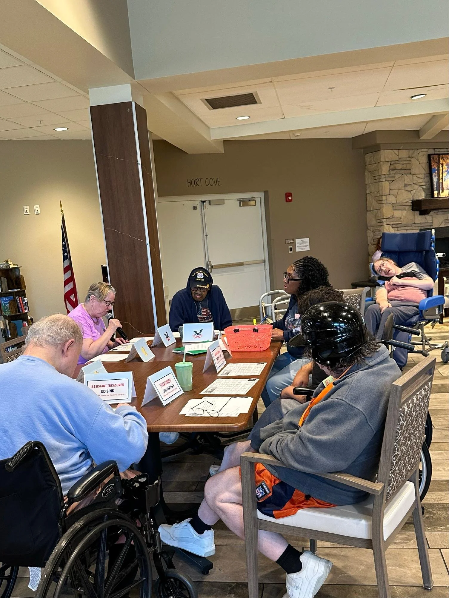 People gathered around a table in a community room, some in wheelchairs, with name tags, papers, and notebooks, in a meeting or discussion setting.